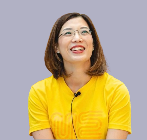 Smiling woman in yellow T-shirt with glasses and earrings, wearing a lapel microphone, at an indoor event with people in the background.