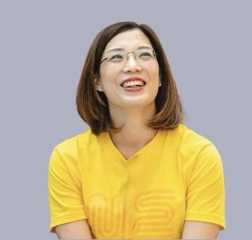 Smiling woman in yellow T-shirt with glasses and earrings, wearing a lapel microphone, at an indoor event with people in the background.