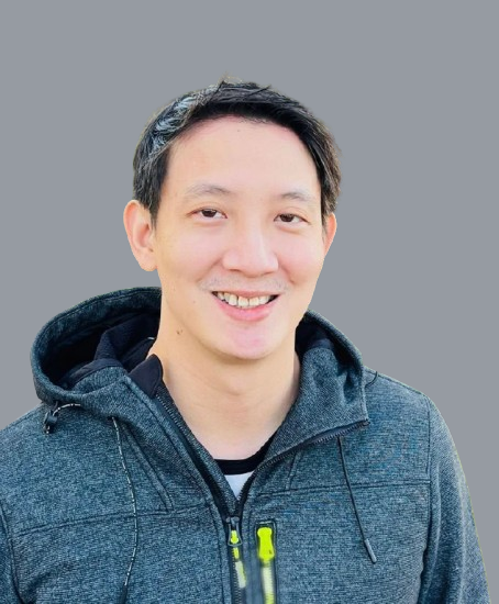 A smiling man with short black hair, wearing a dark gray hoodie with bright yellow zippers, standing outdoors in front of a green hedge.
