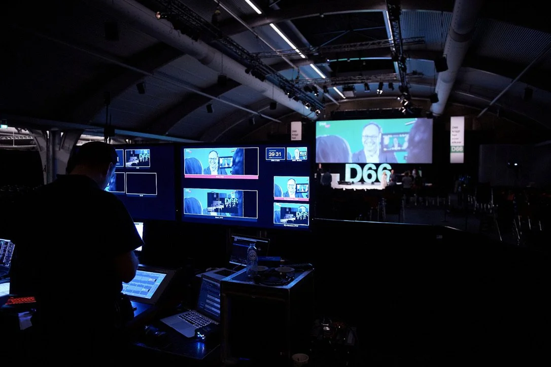 Een persoon die achter een controlepaneel zit in een donkere zaal tijdens een evenement of conference, met meerdere schermen waarop een man in een groen scherm te zien is.