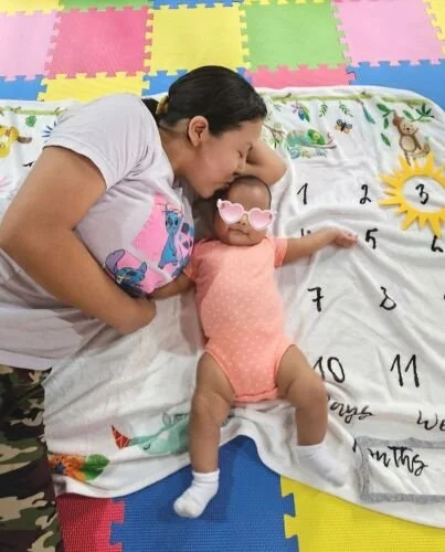 A woman with dark hair and a white t-shirt lying on her side on a multicolored foam mat, next to a baby in a peach onesie with pink sunglasses, lying on a white blanket with numbers and a yellow sun design, reaching out with one arm.