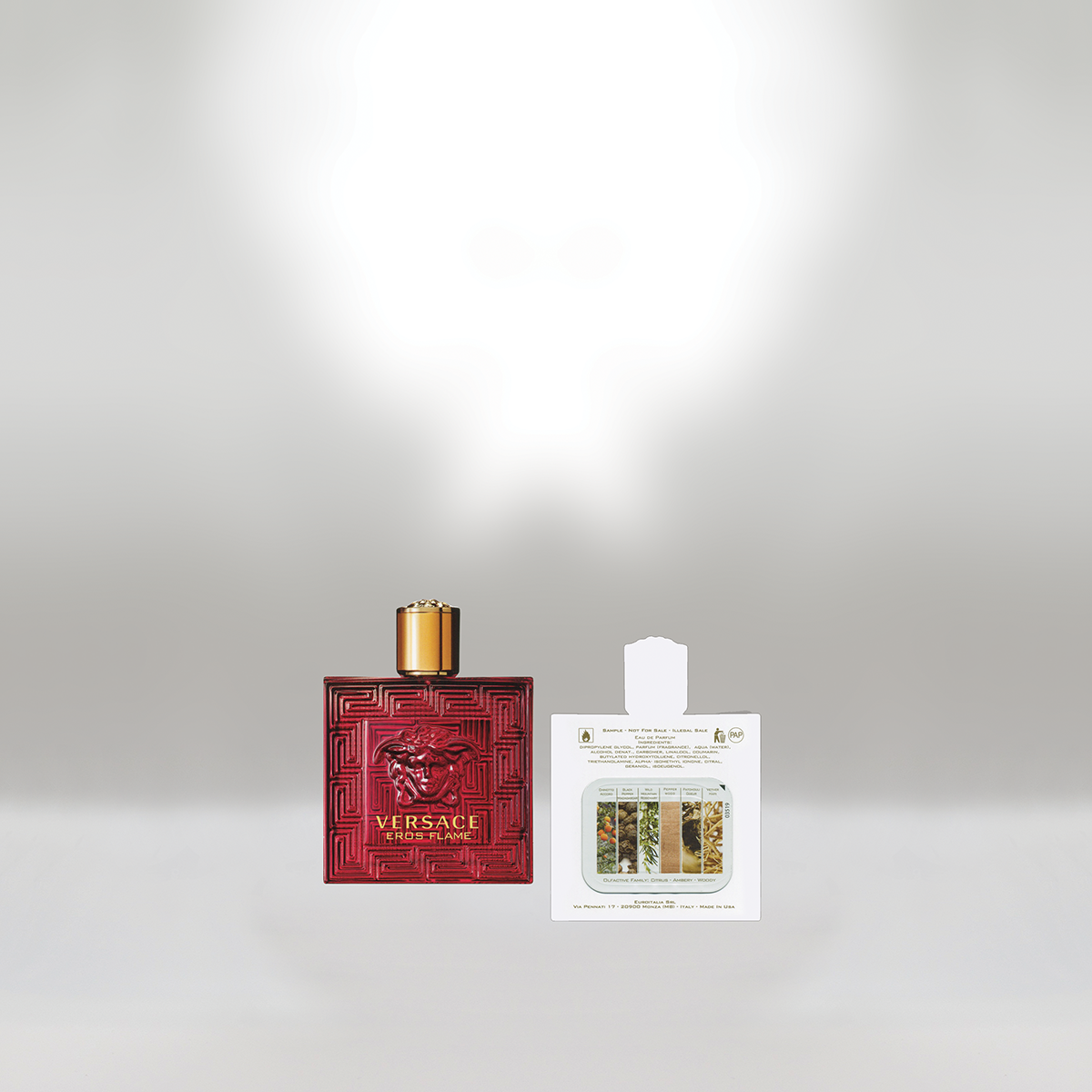 A red Versace Eros Flame perfume bottle with a gold cap next to a sample perfume packet against a plain light gray background.