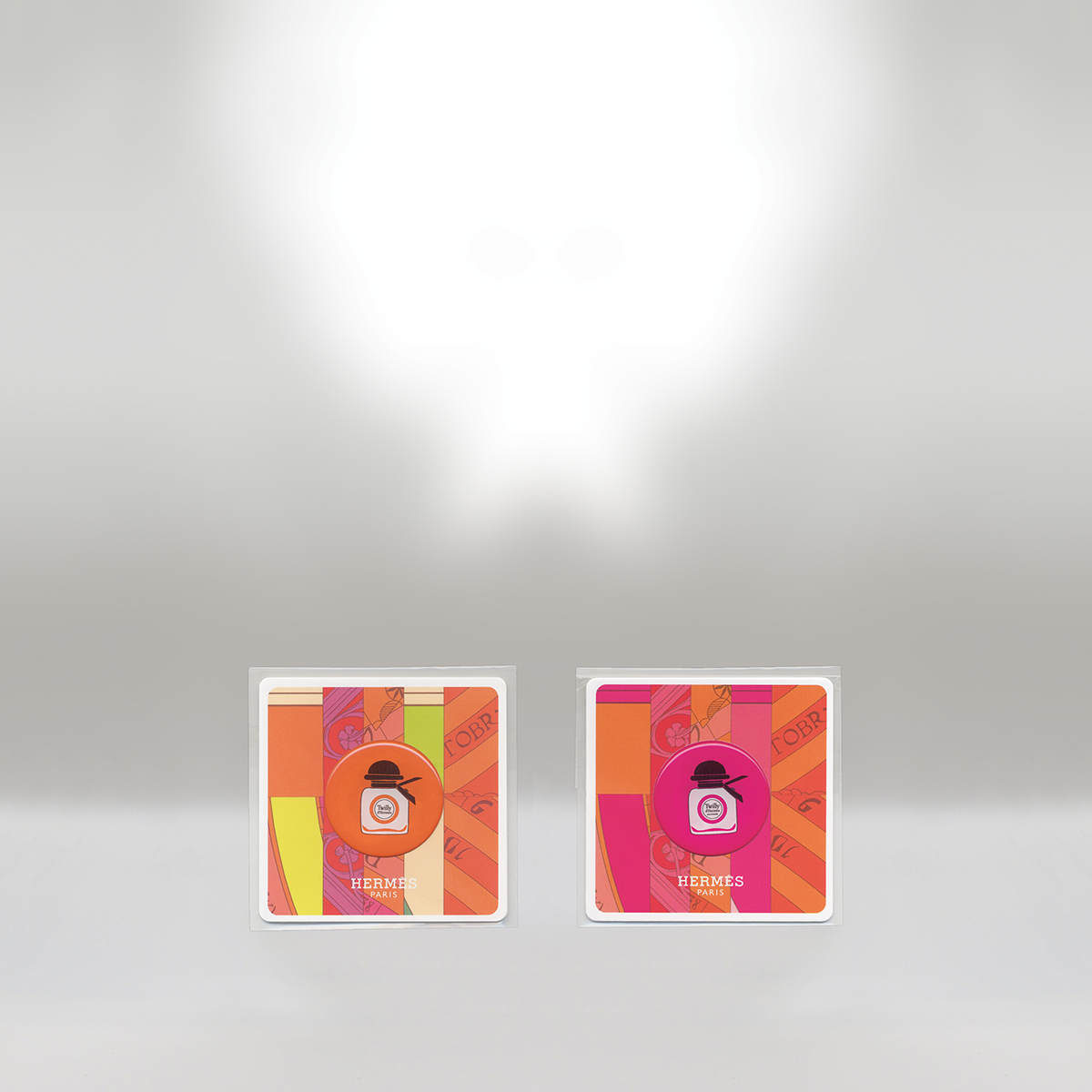 Two Hermes perfume sample cards with colorful abstract patterns, one in orange and the other in pink, placed on a gray surface.