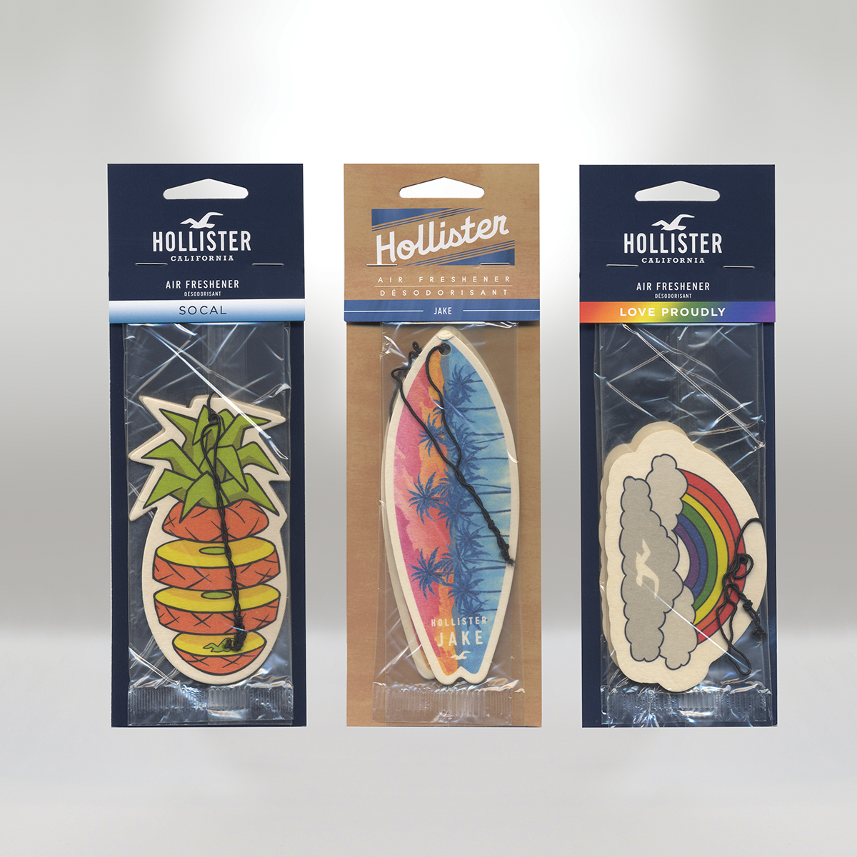 Three Hollister air fresheners in packaging with colorful designs: a pineapple with green leaves, a surfboard with a sunset and palm trees, and a cloud with a rainbow.