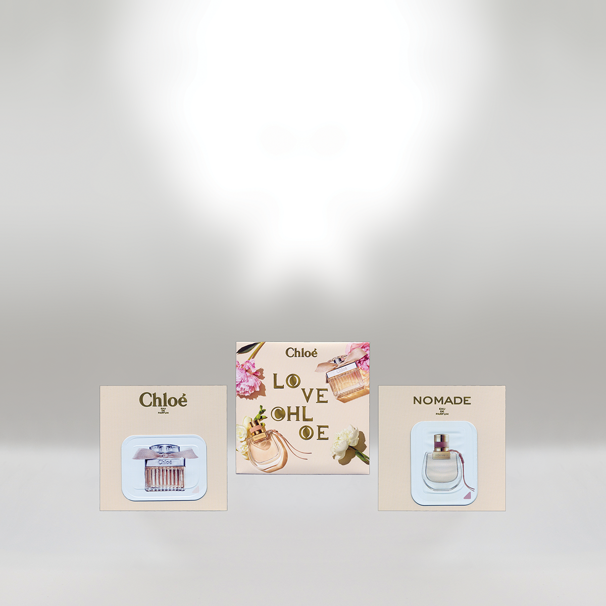 Three perfume bottles on display, with two cardboard backings and a colorful card in the middle. The left bottle on a beige backing labeled 'Chloé', the right bottle on a beige backing labeled 'NOMADE', and the middle card has 'Chloé LOVE CHLOE' with floral and perfume images.