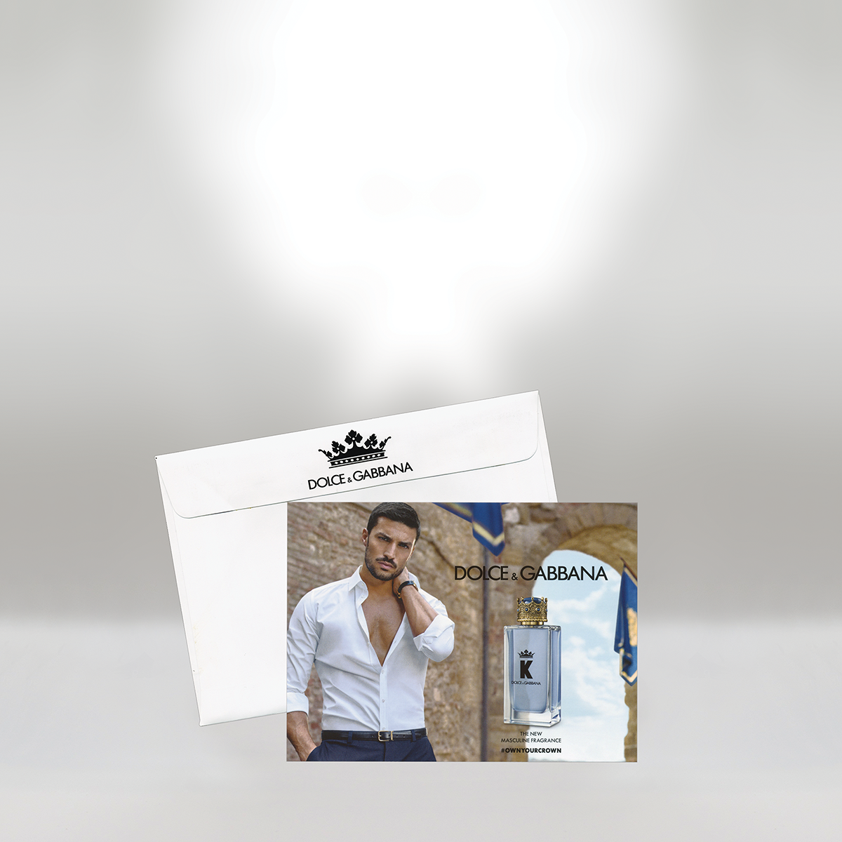 Publicité pour le parfum Dolce & Gabbana représentant un homme en chemise blanche, avec un flacon de parfum et le logo Dolce & Gabbana sur la carte, et une enveloppe avec un logo en forme de couronne et le nom de la marque.