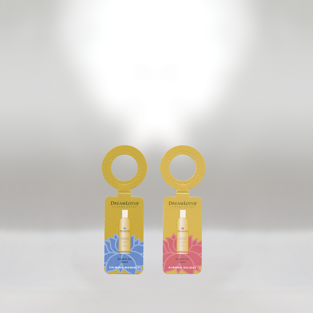 Two Dream Lotus by Kama Sutra bottles with packaging tags, one labeled 'Calming Moments' in blue and the other 'Almond Delight' in pink, hanging from a gold ring, against a plain background.