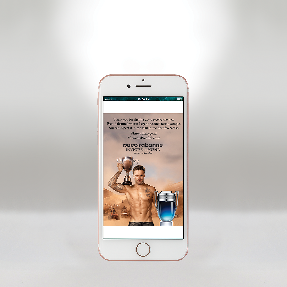 iPhone affichant un message promotionnel de Paco Rabanne Invictus Legend avec un modèle masculin torse nu tenant un grand trophée et se tenant dans un paysage désertique avec des camions en arrière-plan.