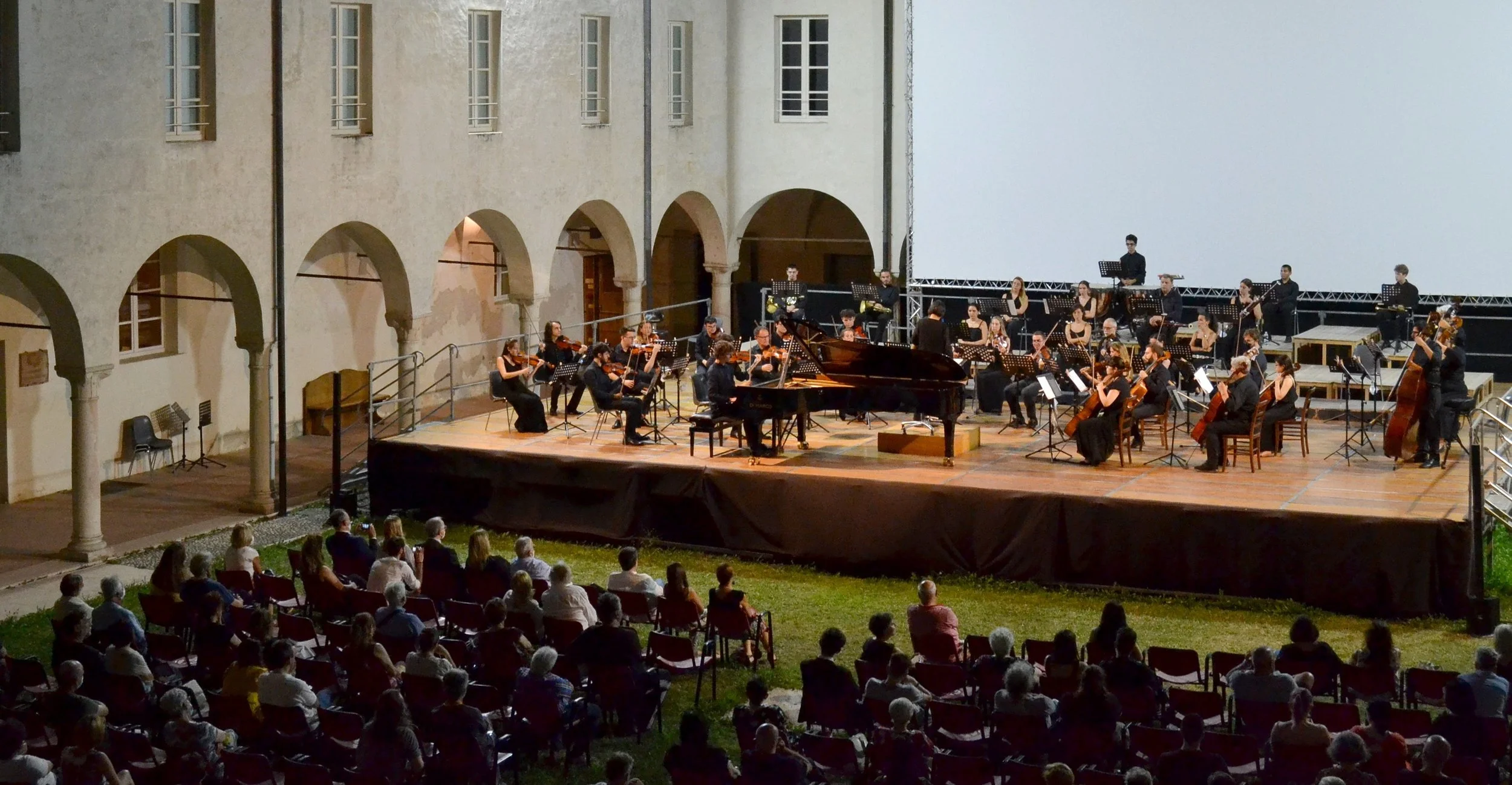 Outdoor orchestra concert with a grand piano and audience seated on grass in a courtyard.