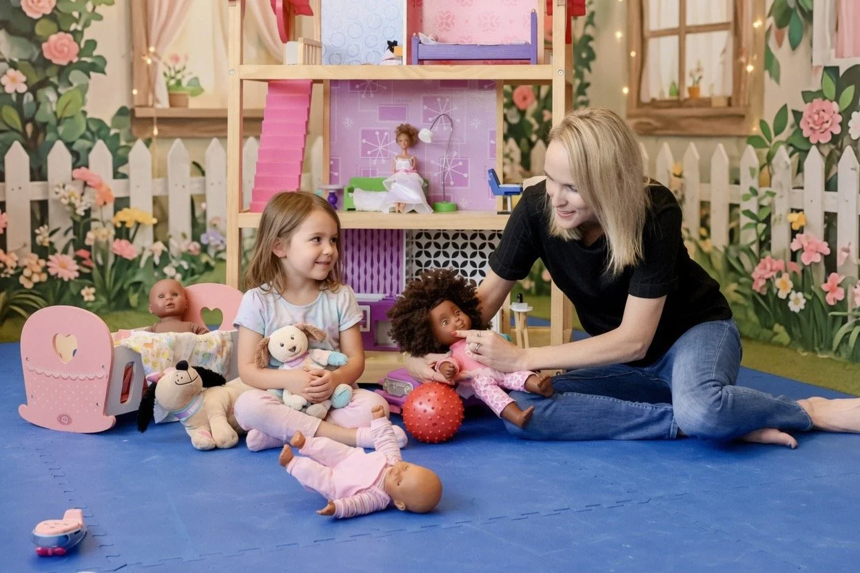 A woman plays with young children and plush toys in a playroom decorated with flowers and fairy lights.