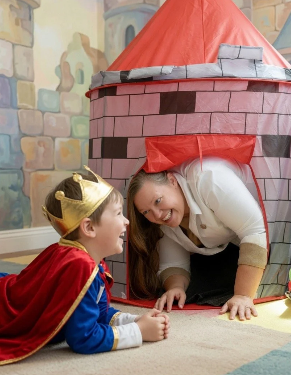 A young boy dressed as a king with a gold crown and red cape is lying on the floor, smiling and looking up at a woman. The woman is kneeling next to a cardboard castle fort, peeking out and smiling back at the boy. The setting appears to be a room with a painted castle mural on the wall.