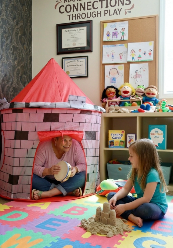 A woman is sitting inside a pink and gray cardboard castle tent, playing a tambourine, with a young girl sitting outside on a colorful alphabet foam mat, playing with sand and a small shovel. The room has a shelf with puppets, children's books, and artwork, and a wall with framed certificates and a quote, "Connections Through Play."