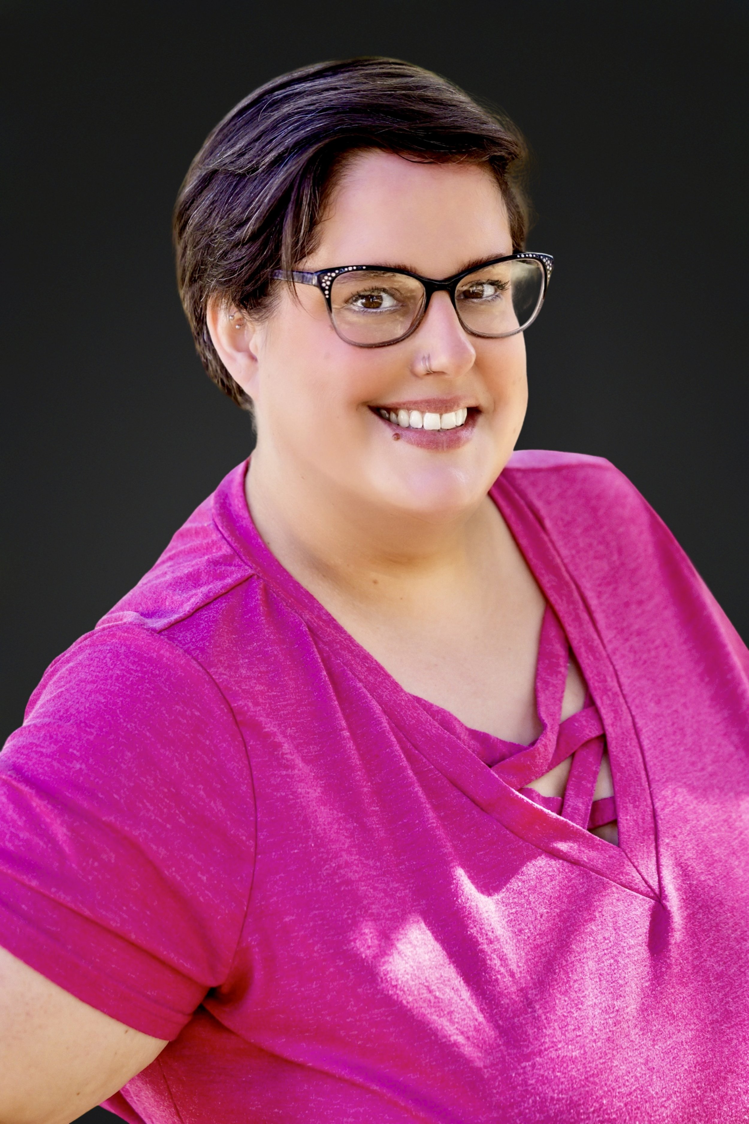 Close-up of a smiling woman with short dark hair, wearing glasses and a pink top, against a black background.