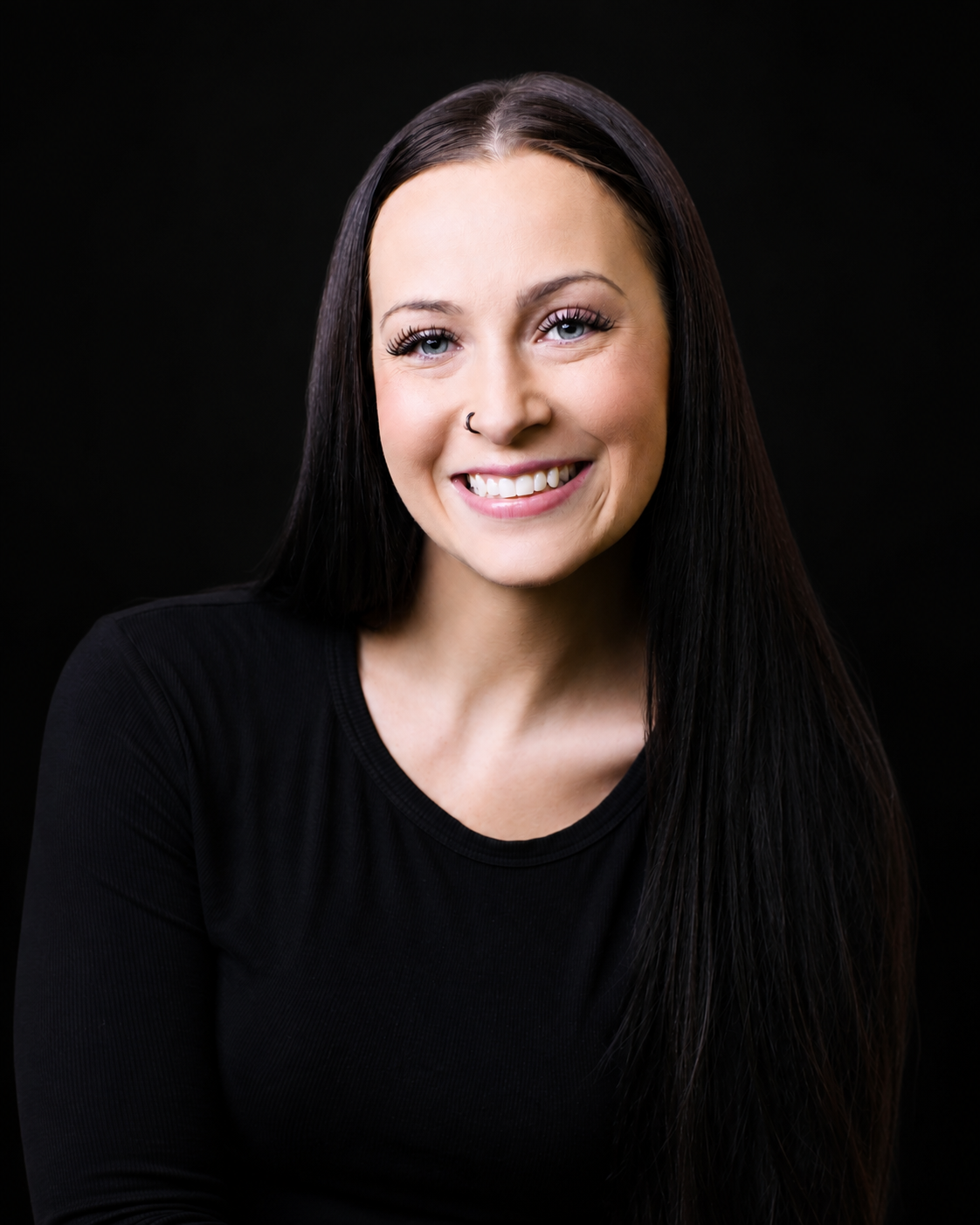 Portrait of a young woman smiling with long black hair, wearing a black top against a black background.