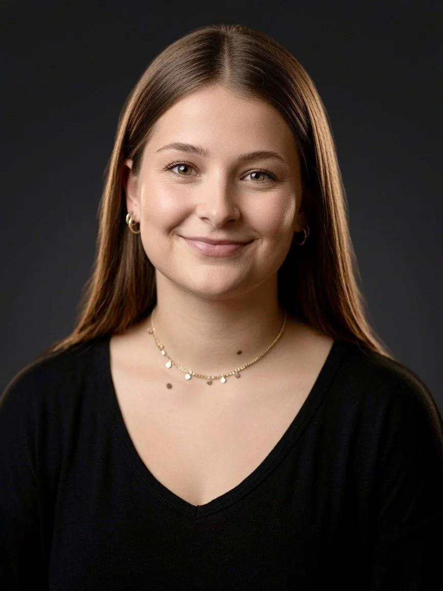 A young smiling woman with long brown hair, wearing a black top, gold necklace, earrings, and a nose piercing, against a dark background.