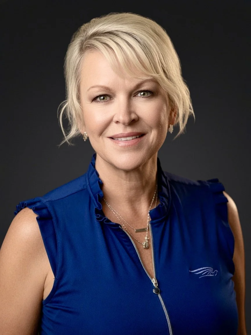 A smiling blonde woman with short hair in a blue sleeveless top, wearing jewelry, against a dark background.