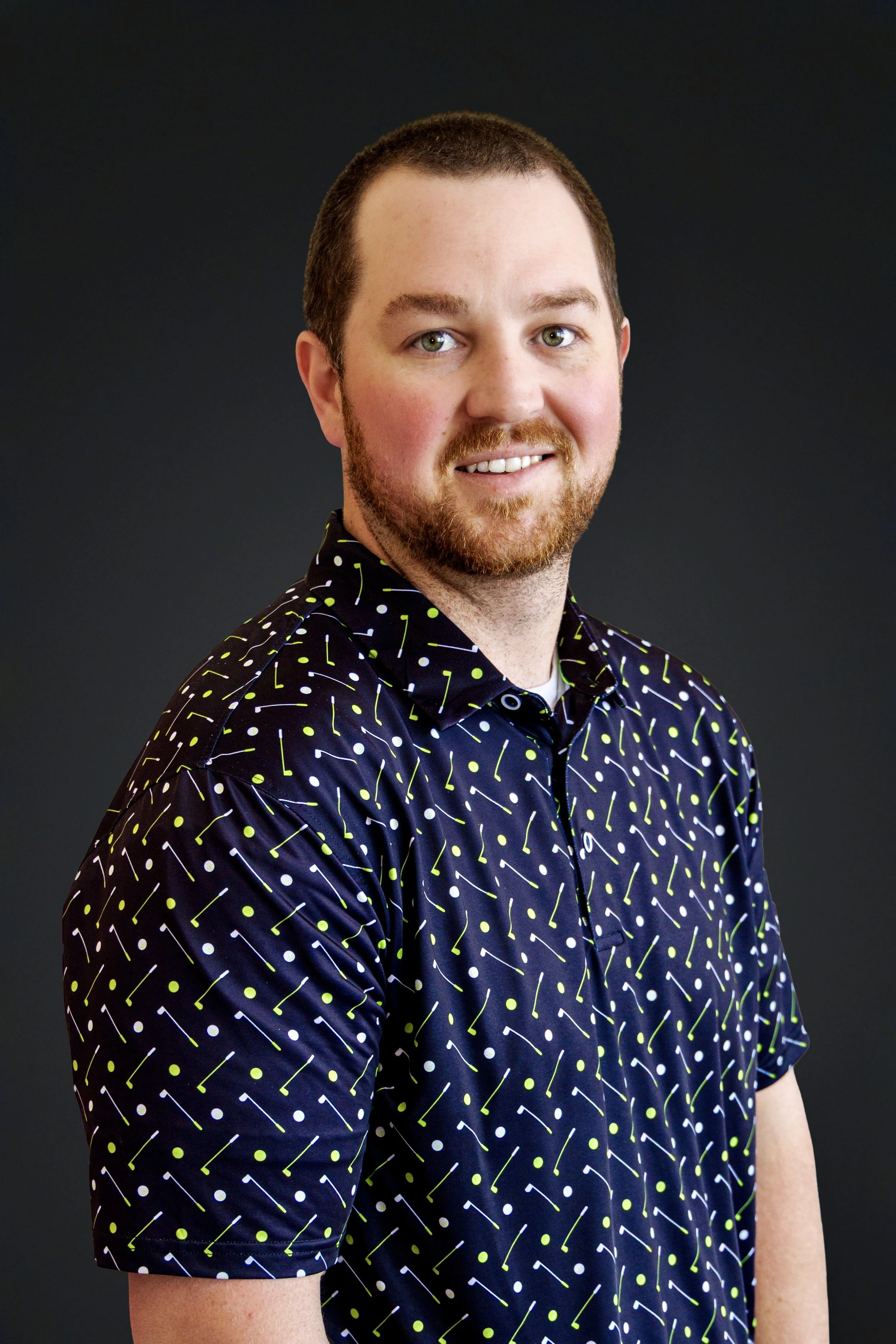 A portrait of a man with short brown hair, a beard, wearing a navy blue shirt with a pattern of yellow and white lines and dots, smiling in front of a black background.