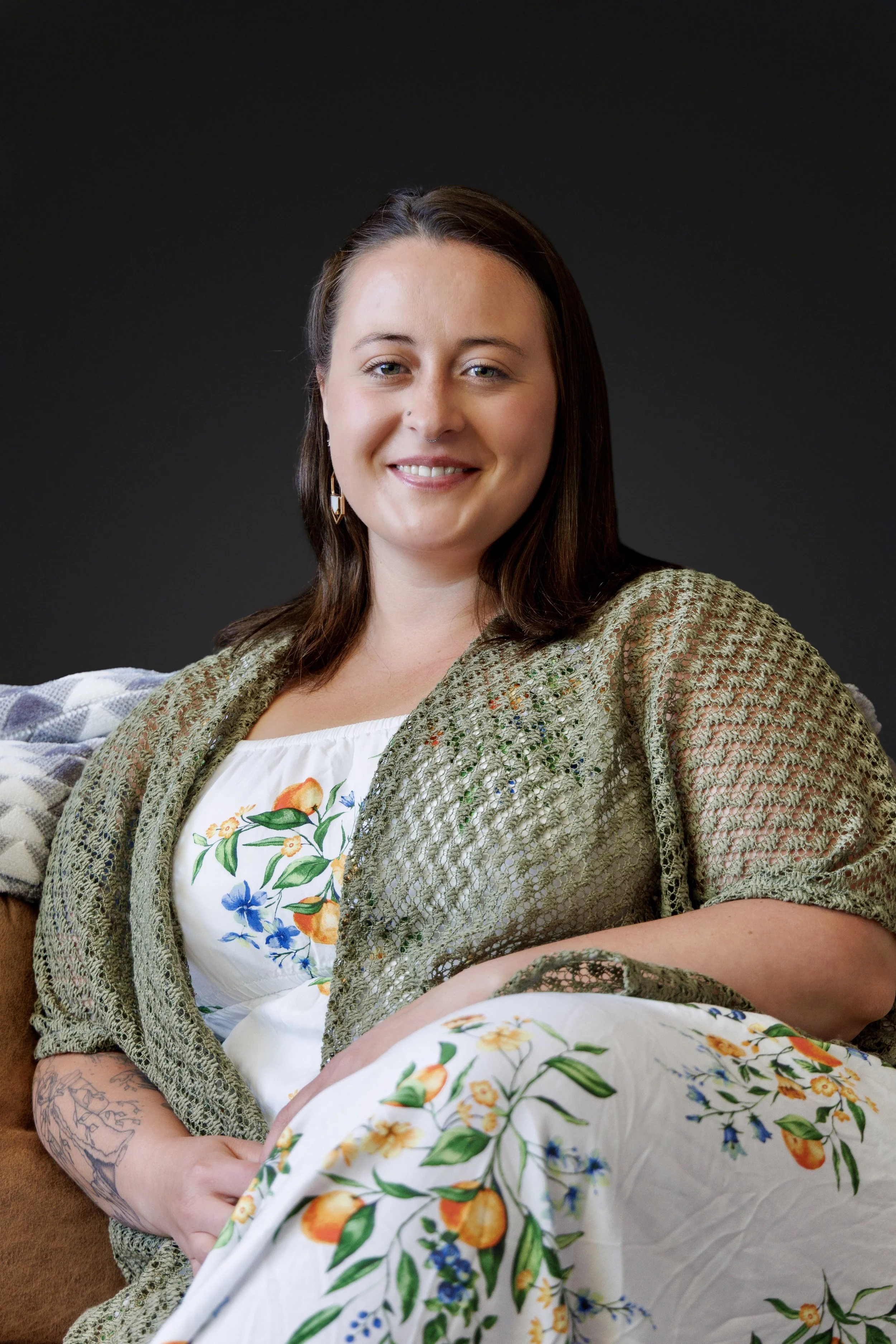 A young woman with brown hair and light skin, smiling, wearing earrings, a green textured cardigan, a white top with a colorful floral print, seated on a couch with a dark background.
