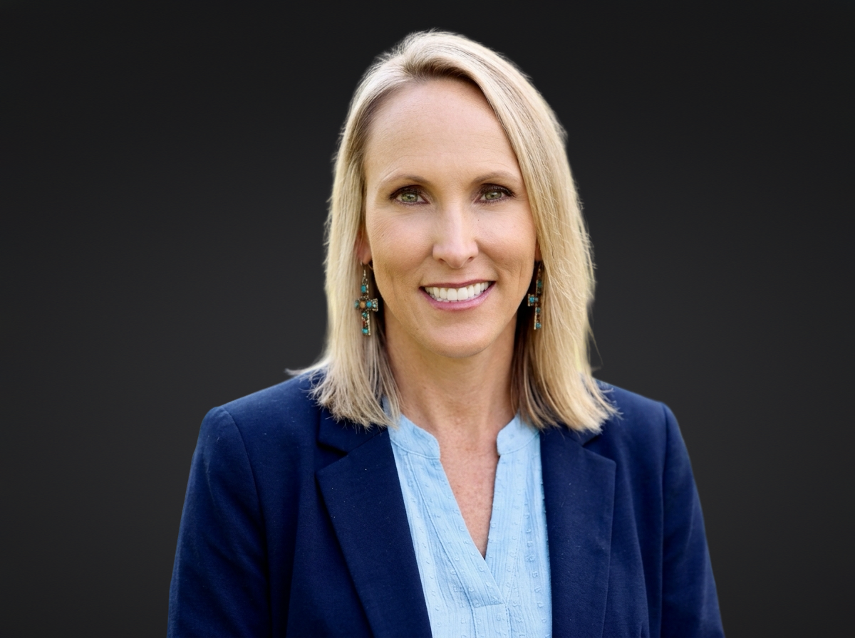 Portrait of a blonde woman smiling, wearing a blue blazer and light blue blouse with turquoise earrings, against a dark background.