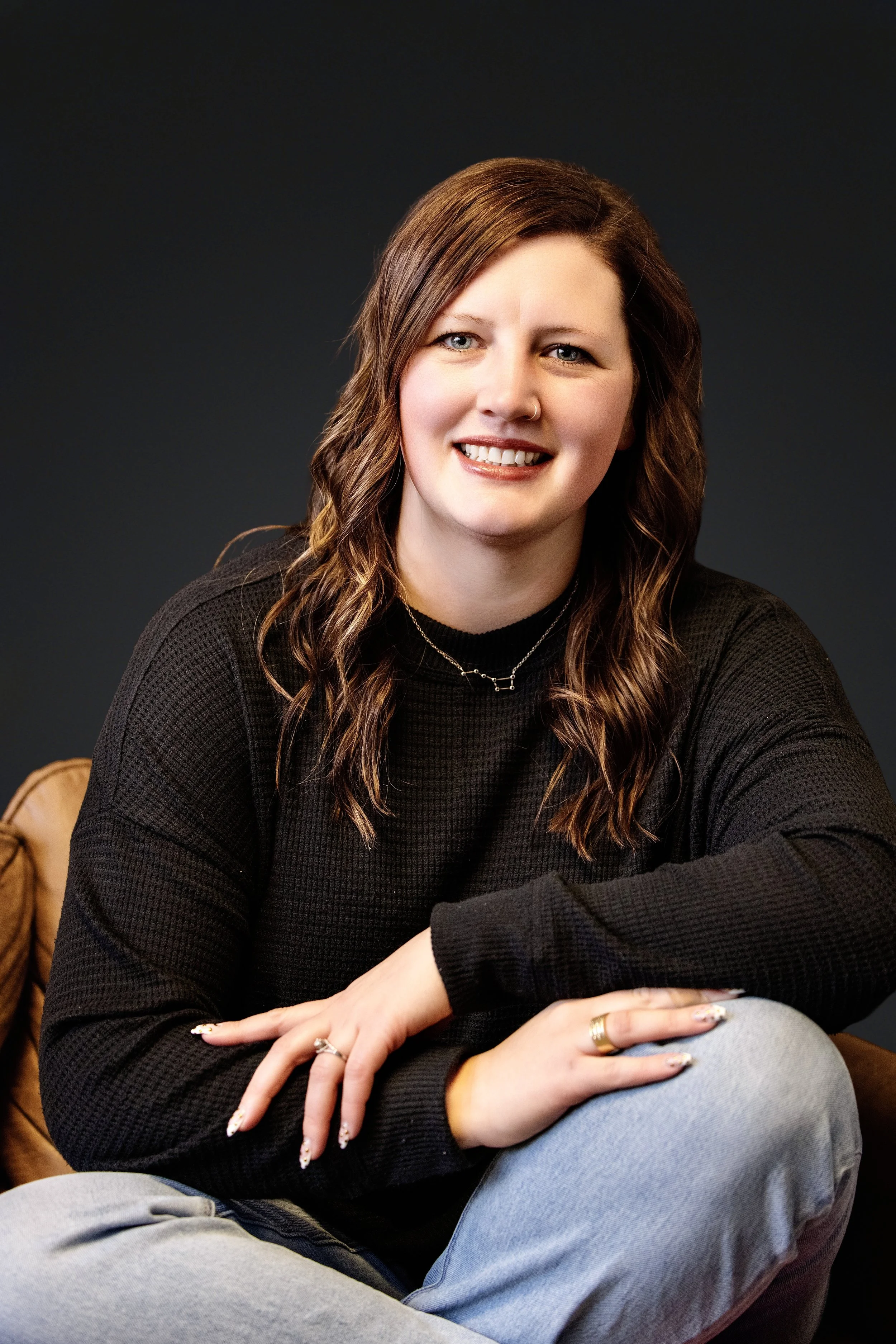 A woman with brown hair and blue eyes smiling, wearing a black sweater and silver necklaces, sitting with crossed legs against a dark background.