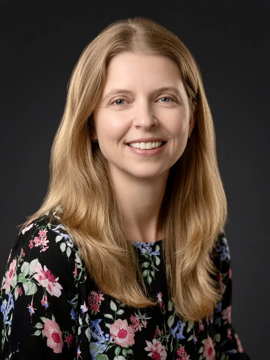 A woman with long, light brown hair smiling, wearing a black floral top, posing against a dark background.