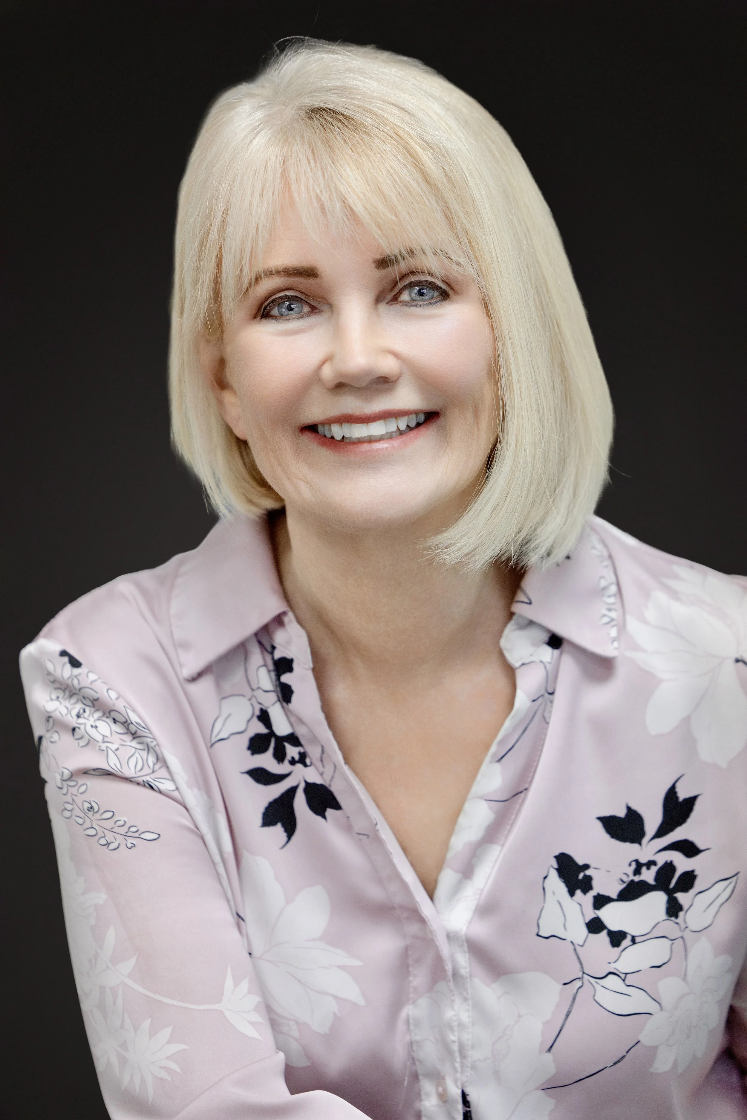 A woman with blonde hair, blue eyes, and a big smile, wearing a light pink shirt with floral patterns, sitting against a dark background.