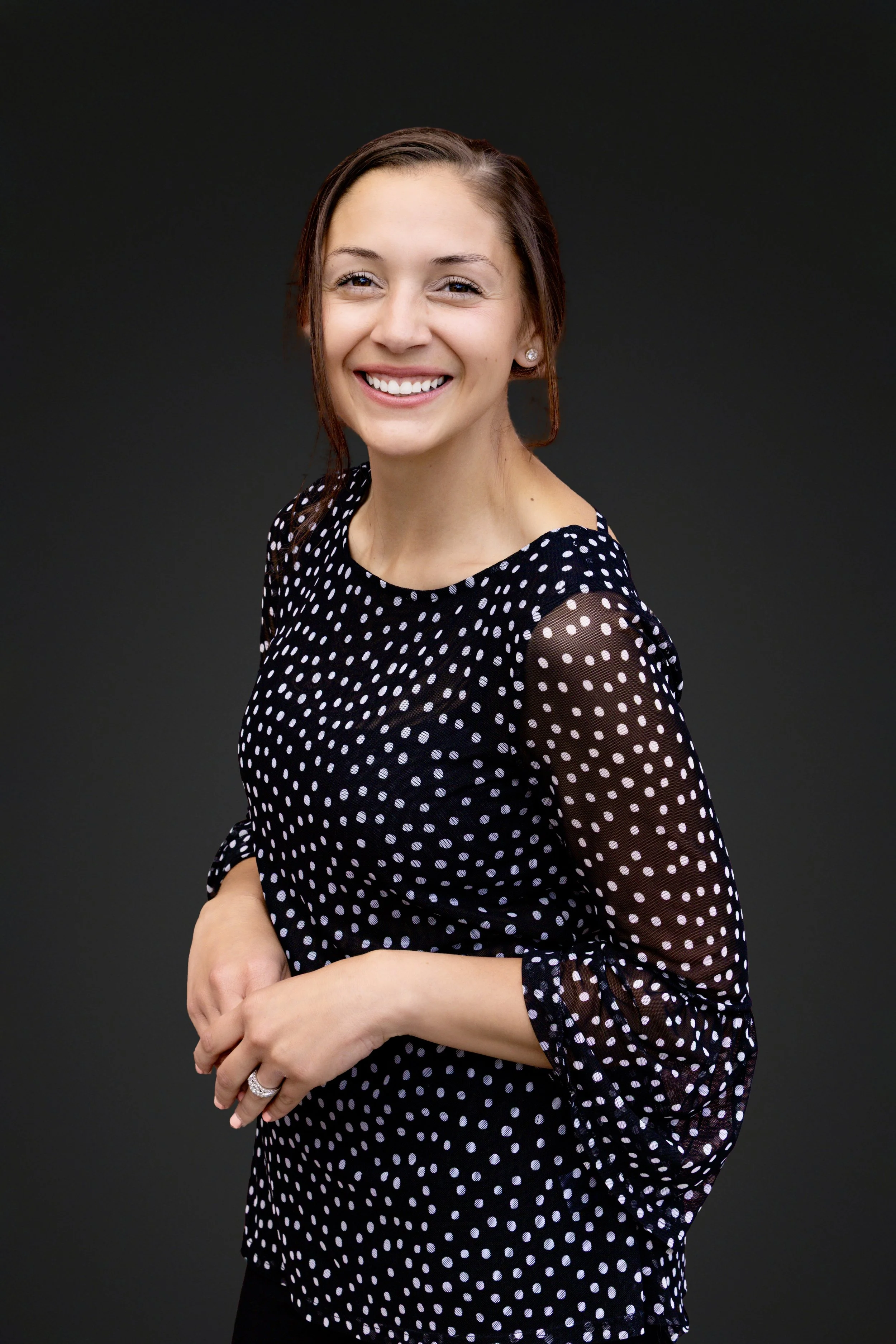 A young woman with long brown hair, smiling, wearing pearl earrings and a black dress with white polka dots, standing against a dark gray background.