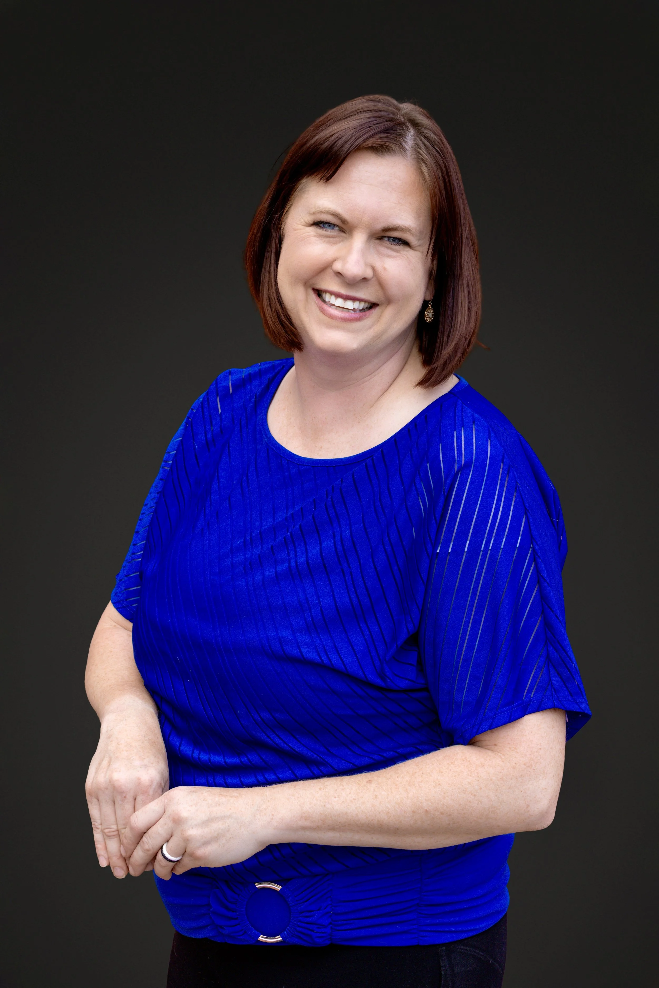 A woman with shoulder-length auburn hair and blue eyes smiling, wearing a bright blue top with vertical lines, standing against a dark background.