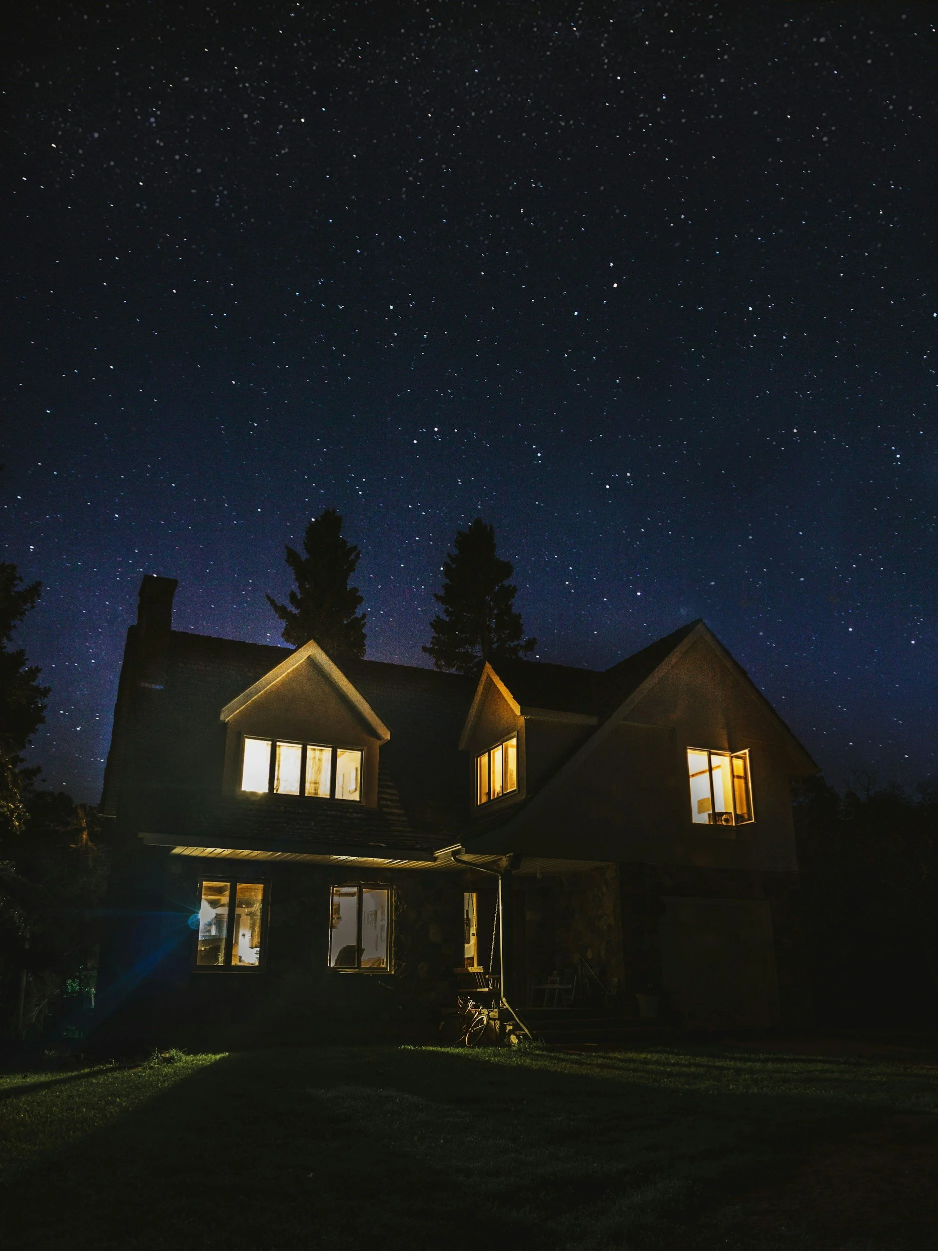A house with several lit windows at night under a starry sky, surrounded by trees and a grassy yard.