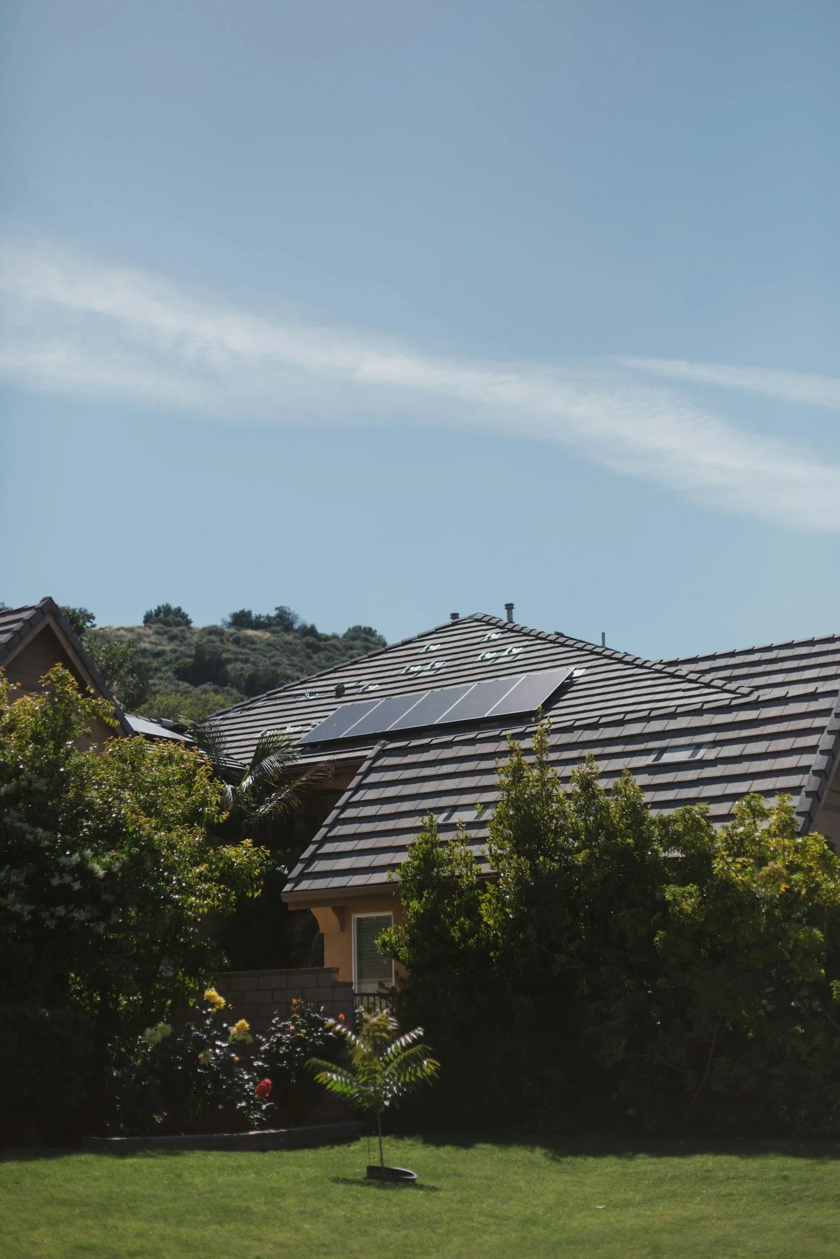 A house with solar panels on its roof, surrounded by green trees and bushes, under a blue sky with wispy clouds.