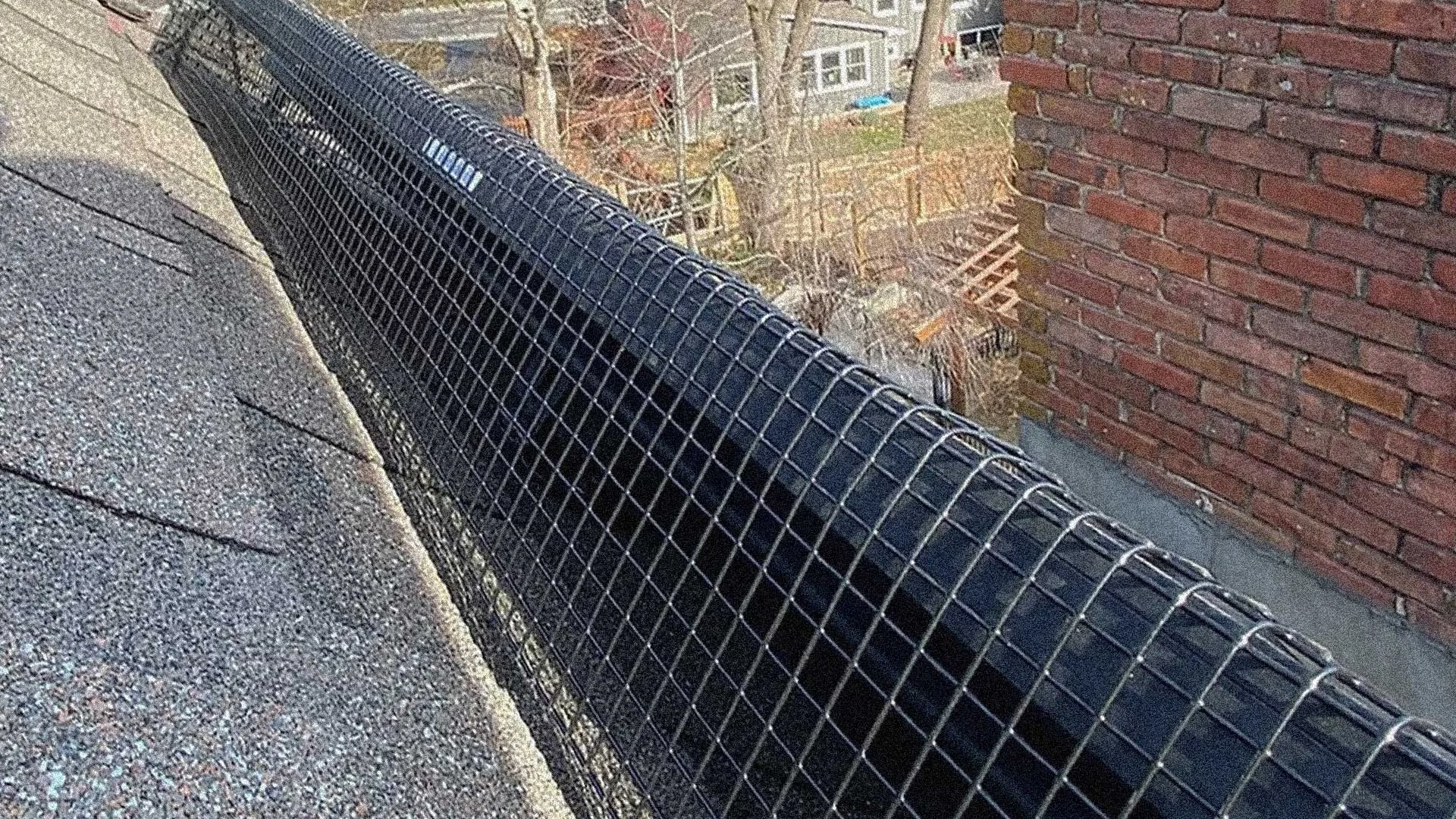 Black drainage pipe on the roof, covered with a wire mesh guard, next to a brick wall with residential buildings and trees in the background.