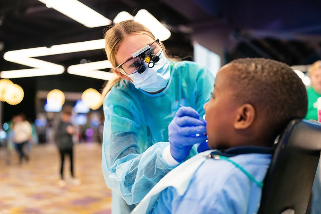 A healthcare worker wearing glasses with magnifying lenses, a face mask, gloves, and a teal medical gown, is examining a young boy's mouth in a brightly lit indoor setting, possibly a clinic or vaccination site.
