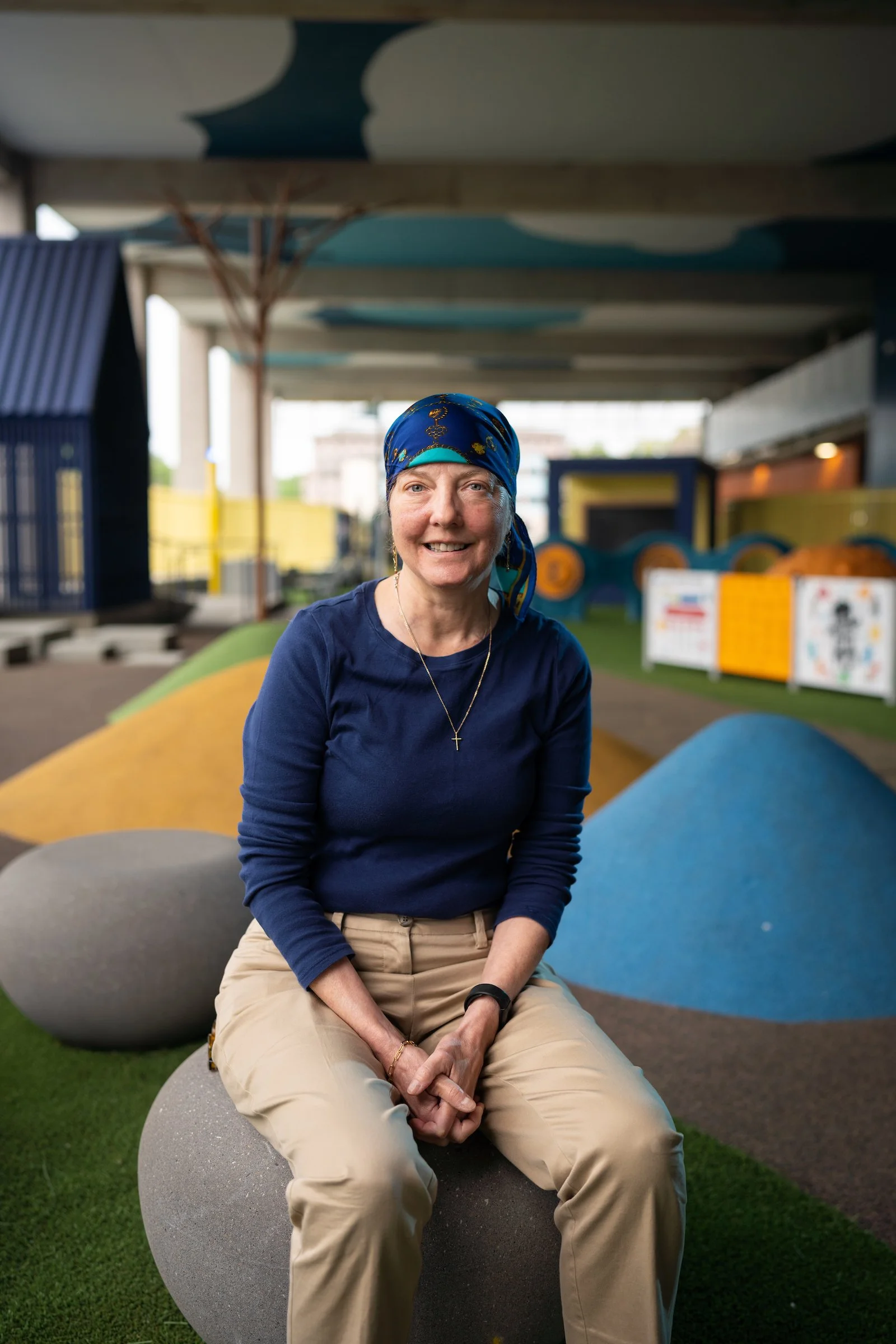 A smiling woman in a blue headscarf and navy blue shirt sitting on a spherical stone bench at a colorful outdoor playground.