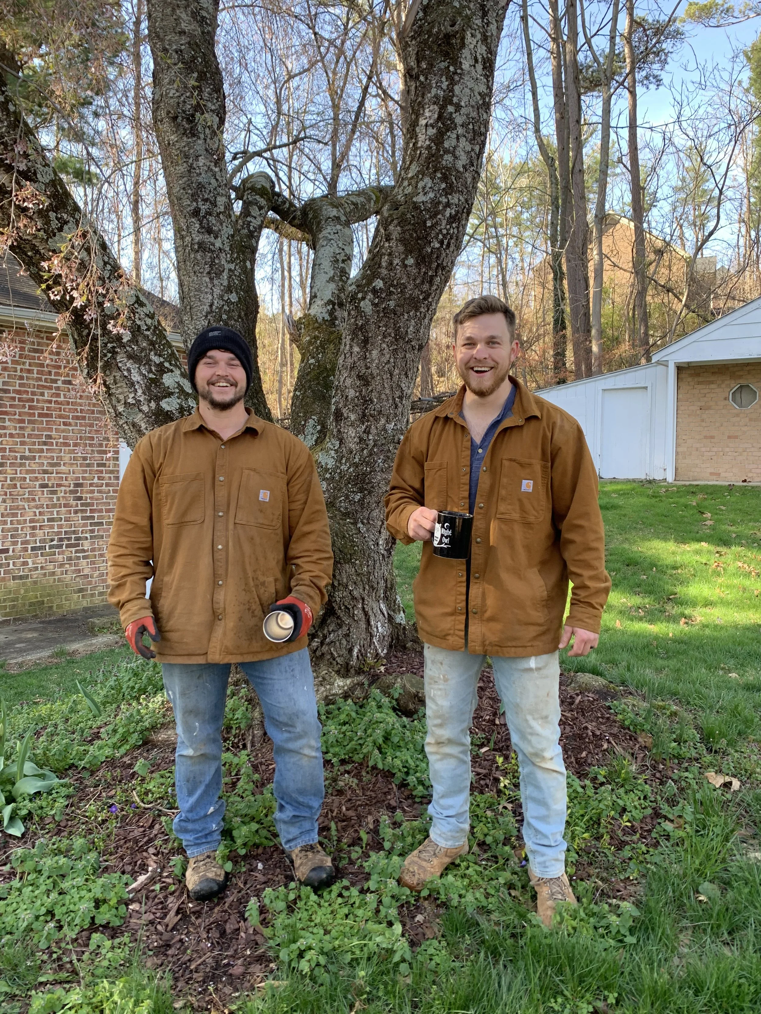 Two men standing outdoors in front of a large tree with a background of suburban houses, smiling and holding gardening tools and a coffee mug, dressed in brown work jackets.