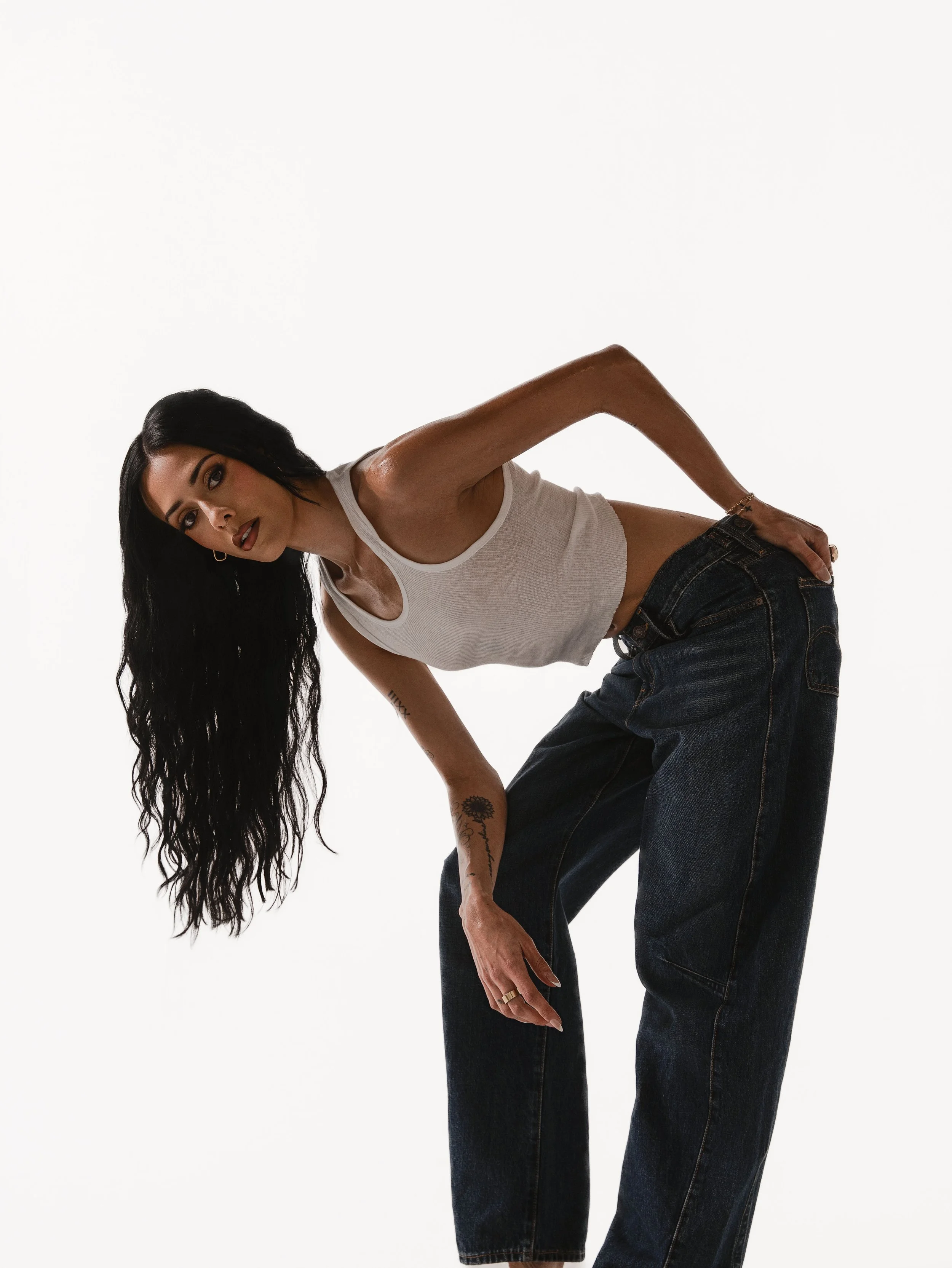 A woman with long black wavy hair wearing a white tank top and dark jeans leaning to the side against a plain white background.