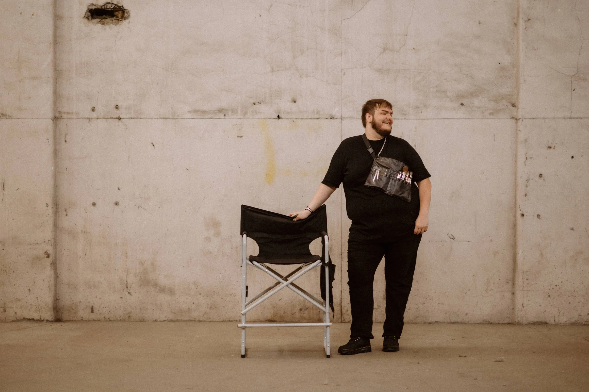 A smiling man with a beard dressed in black, standing indoors against a concrete wall, leaning on a black folding chair, wearing a tool pouch with makeup brushes.