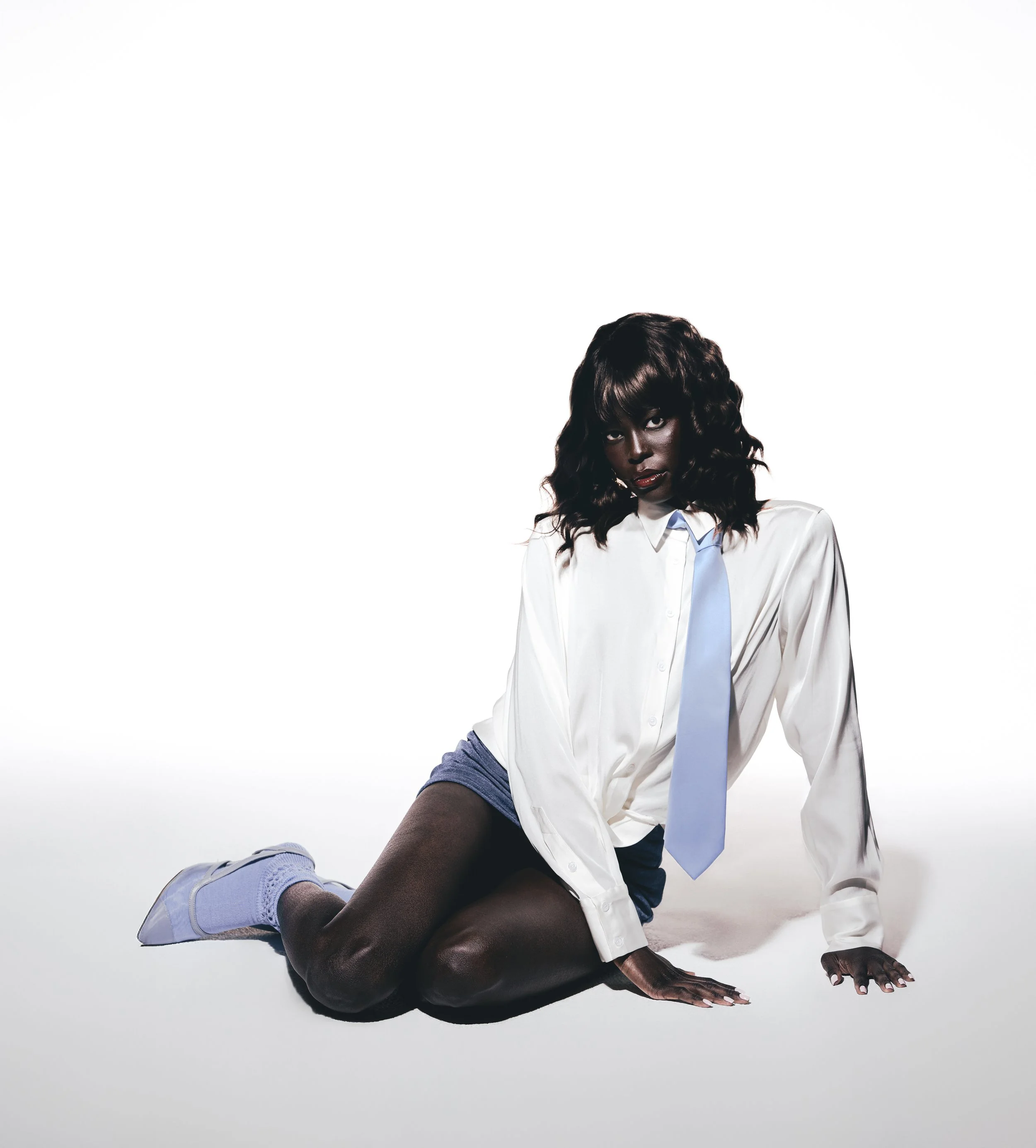 A woman with dark skin and curly hair wearing a white shirt, blue tie, black shorts, and purple sneakers, sitting on the floor against a plain white background.