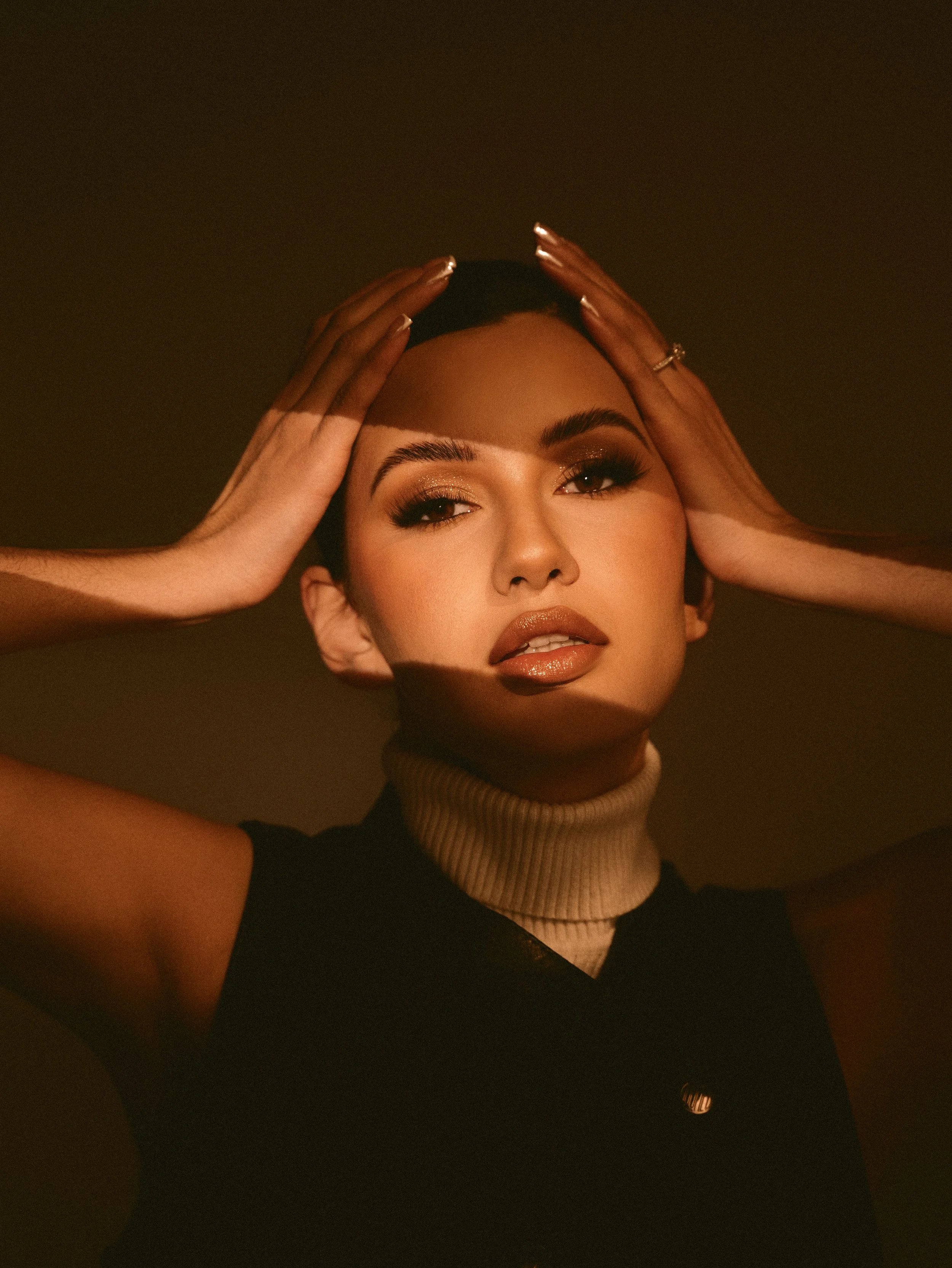 A woman with makeup and a turtleneck poses with hands on her head in warm lighting.