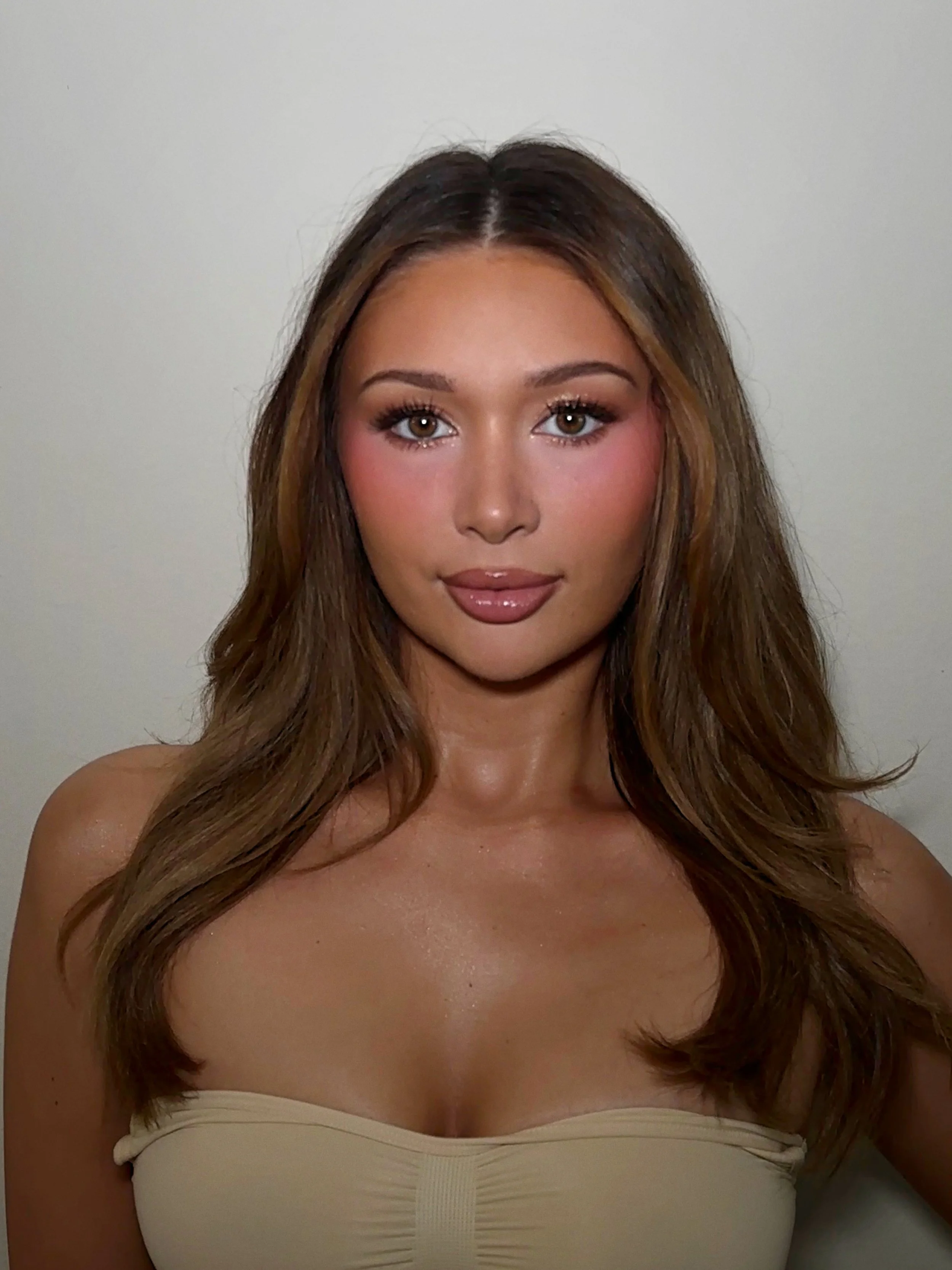 A young woman with long brown hair and light makeup, wearing a beige strapless top against a plain white wall.