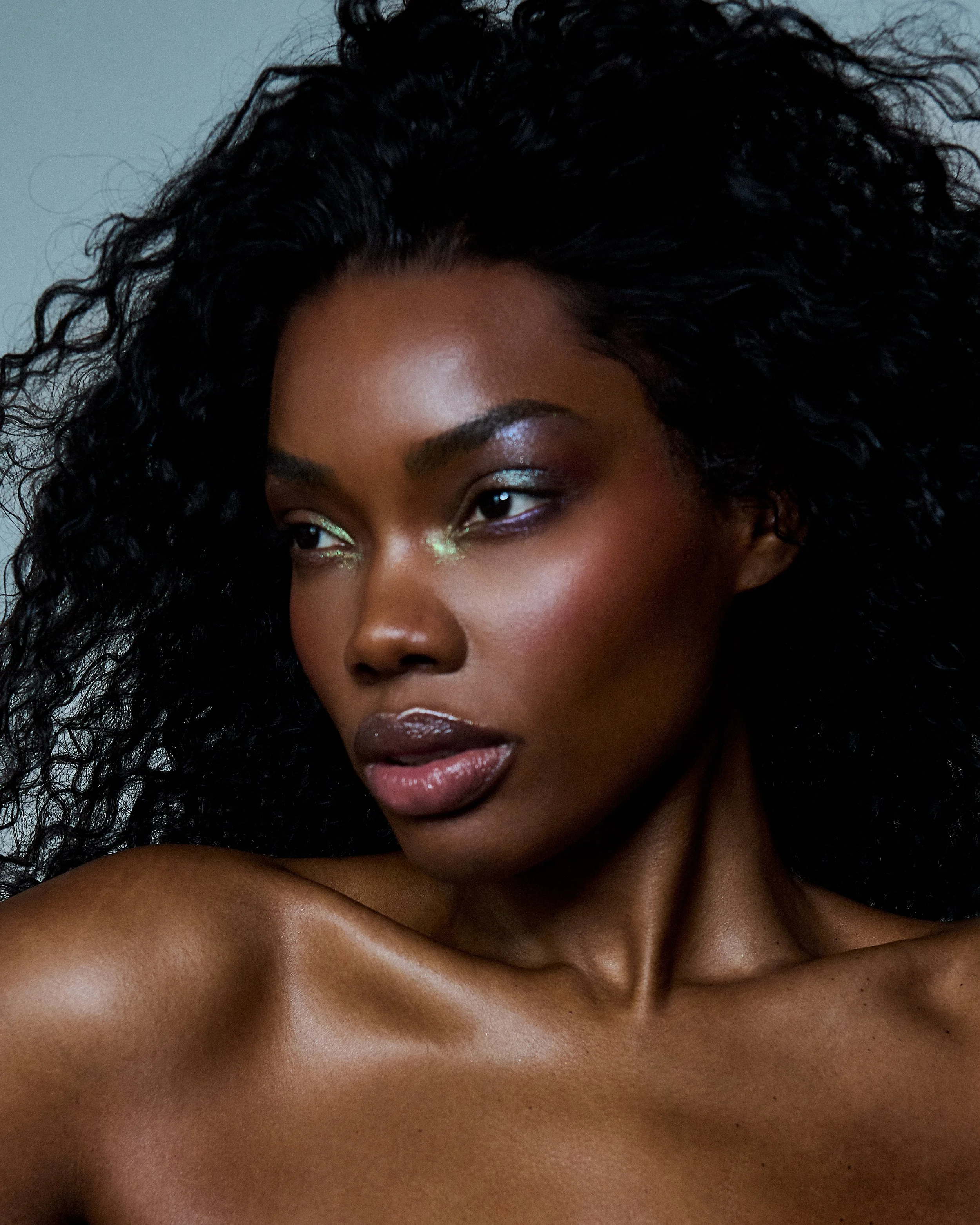 Close-up portrait of a woman with dark skin and curly black hair, wearing shiny eye makeup and glossy lipstick.