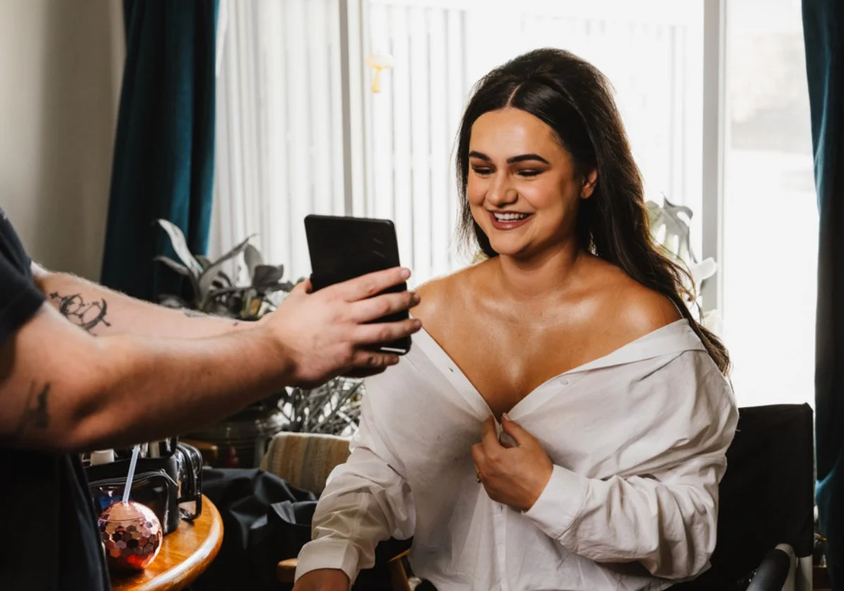 A woman with long brunette hair, smiling, partially undressing, while a tattooed person shows her a phone and takes a photo in a room with natural light and blue curtains.