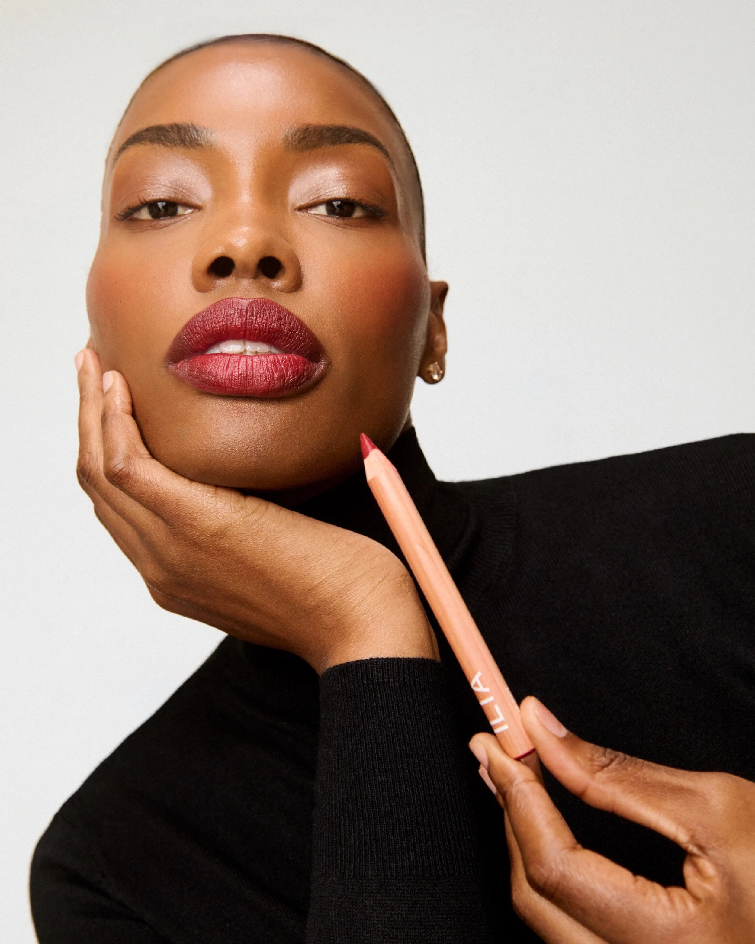 A woman with dark skin and a shaved head holding a pink lip liner pencil near her chin, wearing a black turtleneck, and posing against a plain white background.