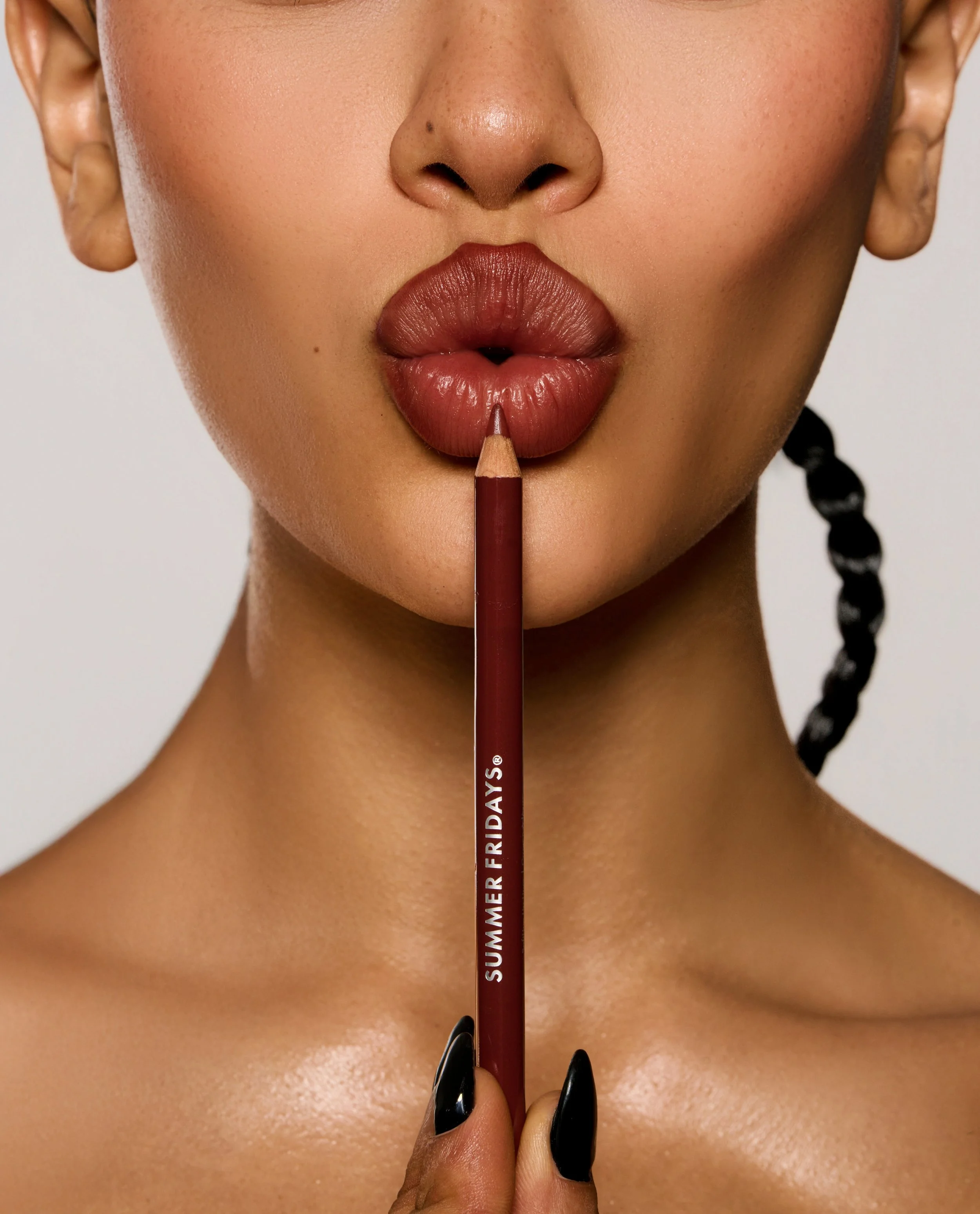 Close-up of a woman with brown lipstick, holding a lip liner pencil labeled 'SUMMER FRIDAYS®' close to her lips, with her hair in a black braid.