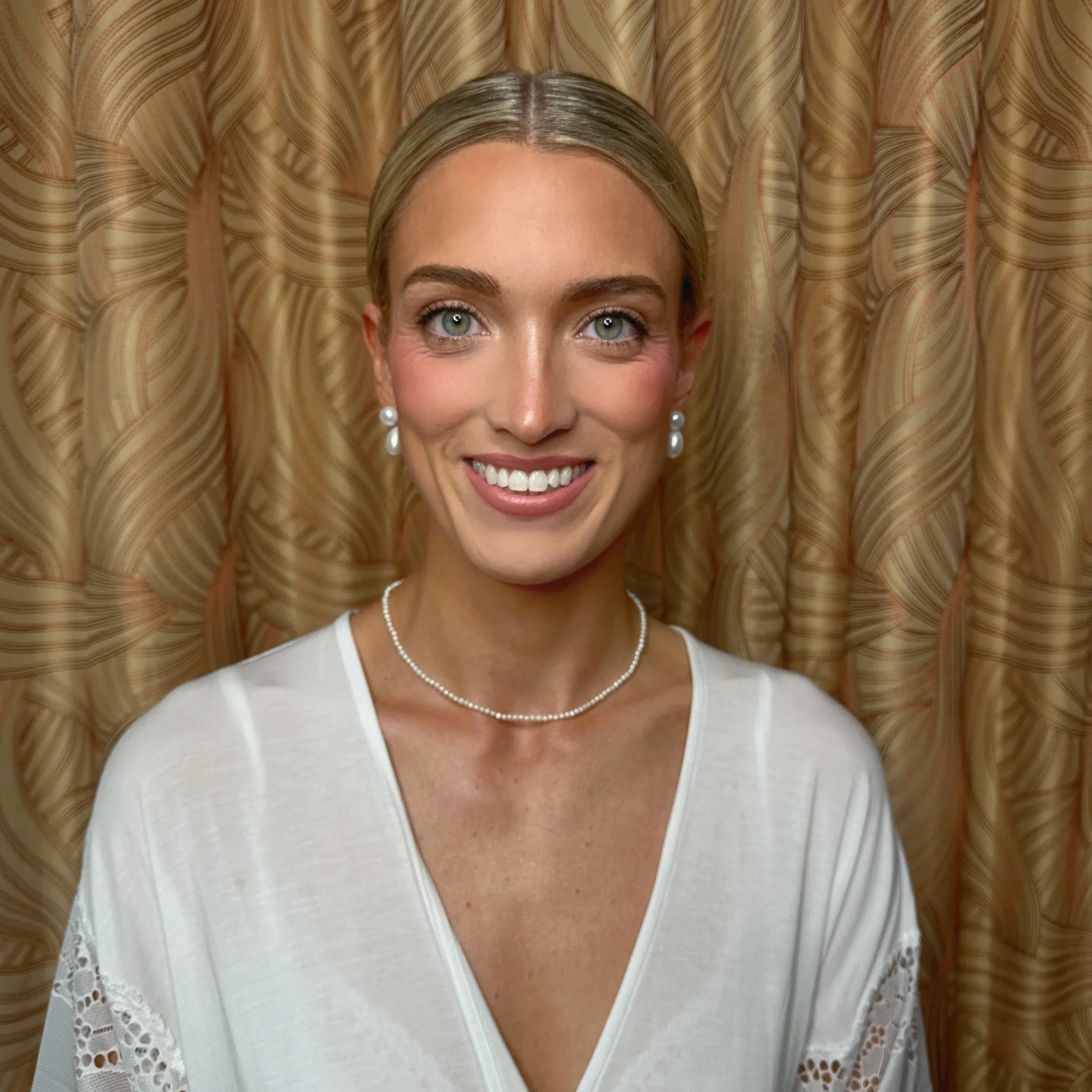 A smiling woman with blonde hair, wearing pearl earrings, a pearl necklace, and a white top, standing in front of a textured, wavy-patterned curtain.