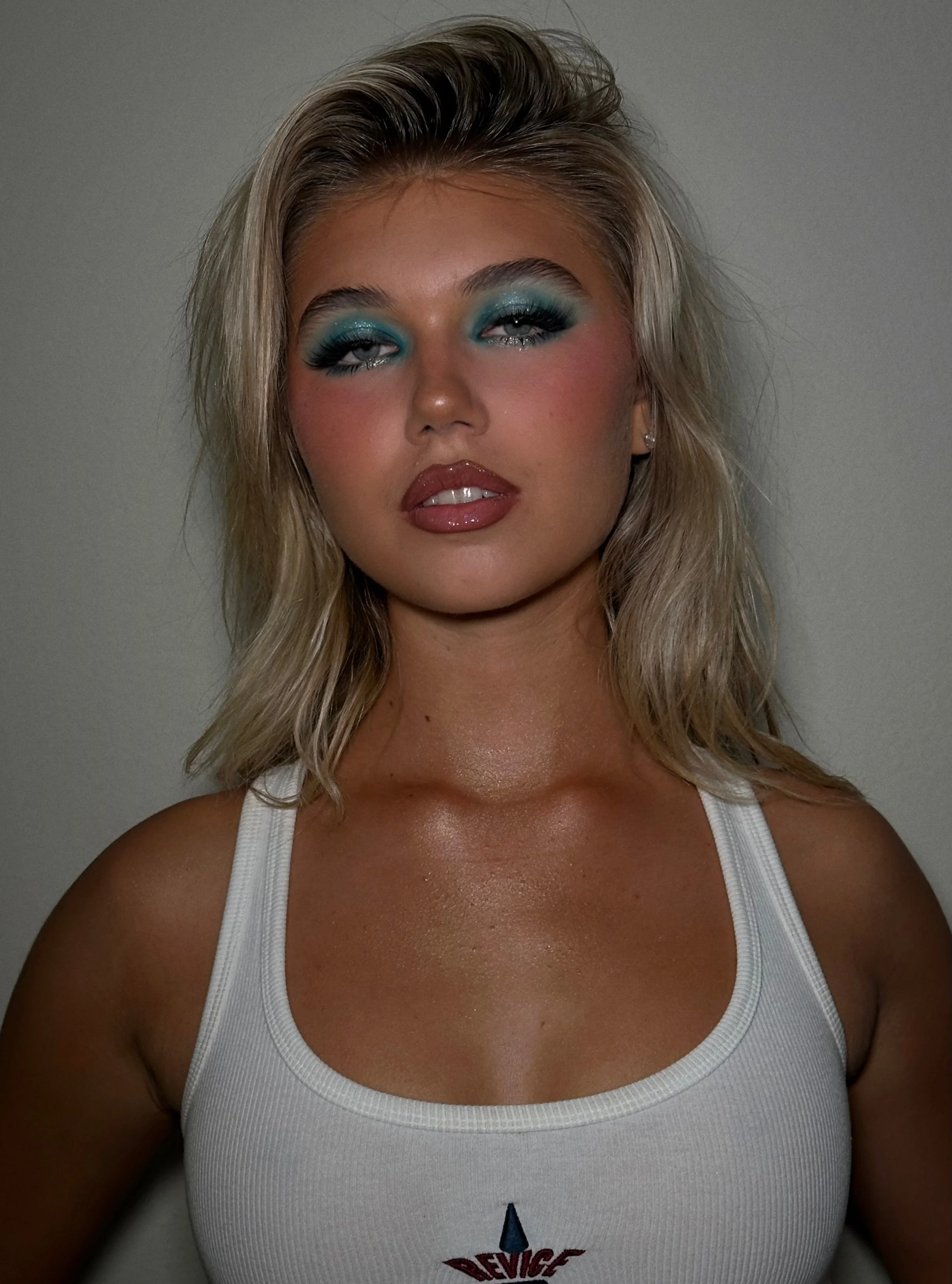 A woman with blonde hair, vibrant blue eye makeup, glossy lips, wearing a white tank top with a logo on it, standing against a plain wall.