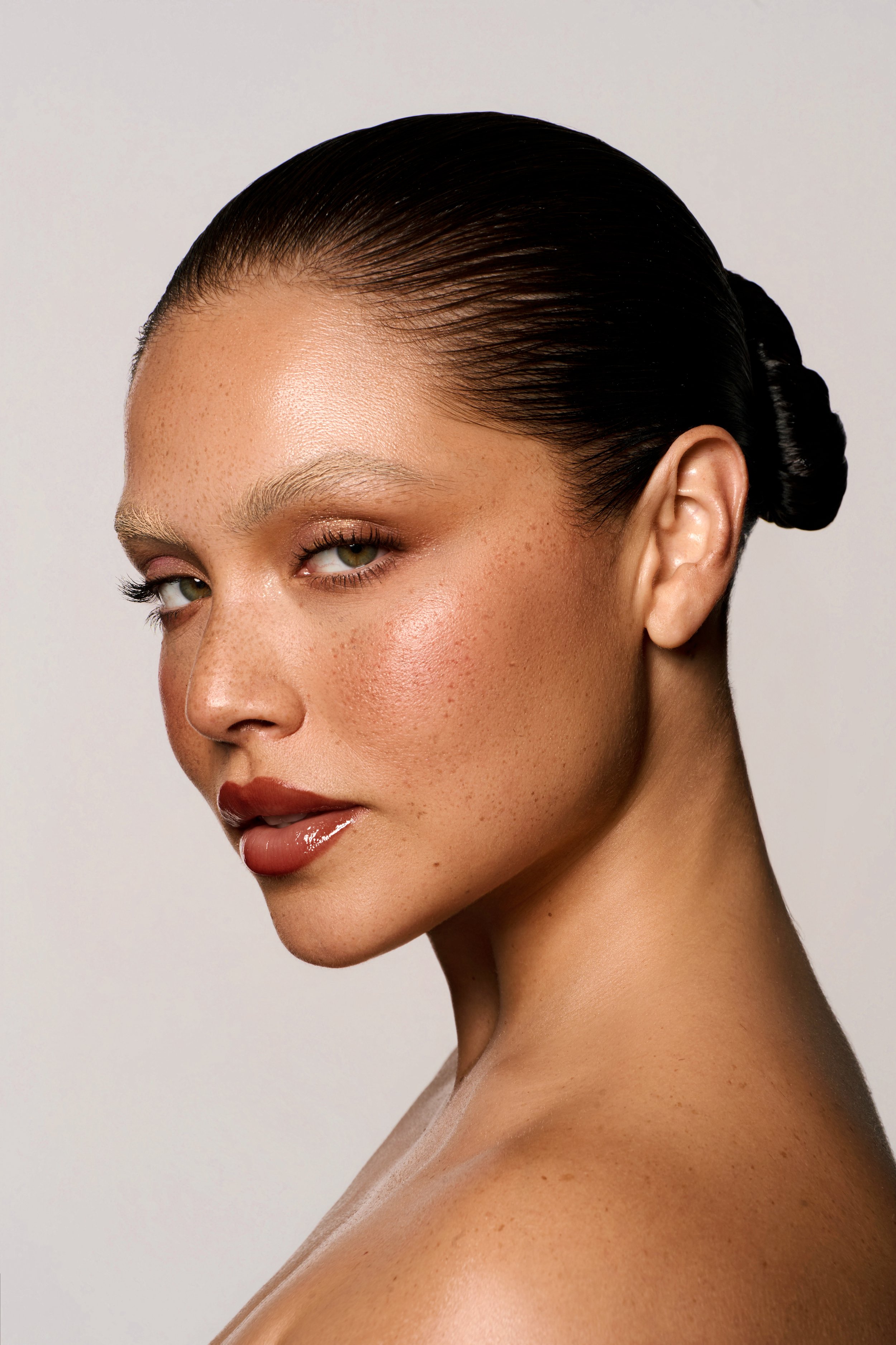 Close-up of a woman with slicked-back dark hair, light skin, and clear complexion, looking over her shoulder with a subtle smile.