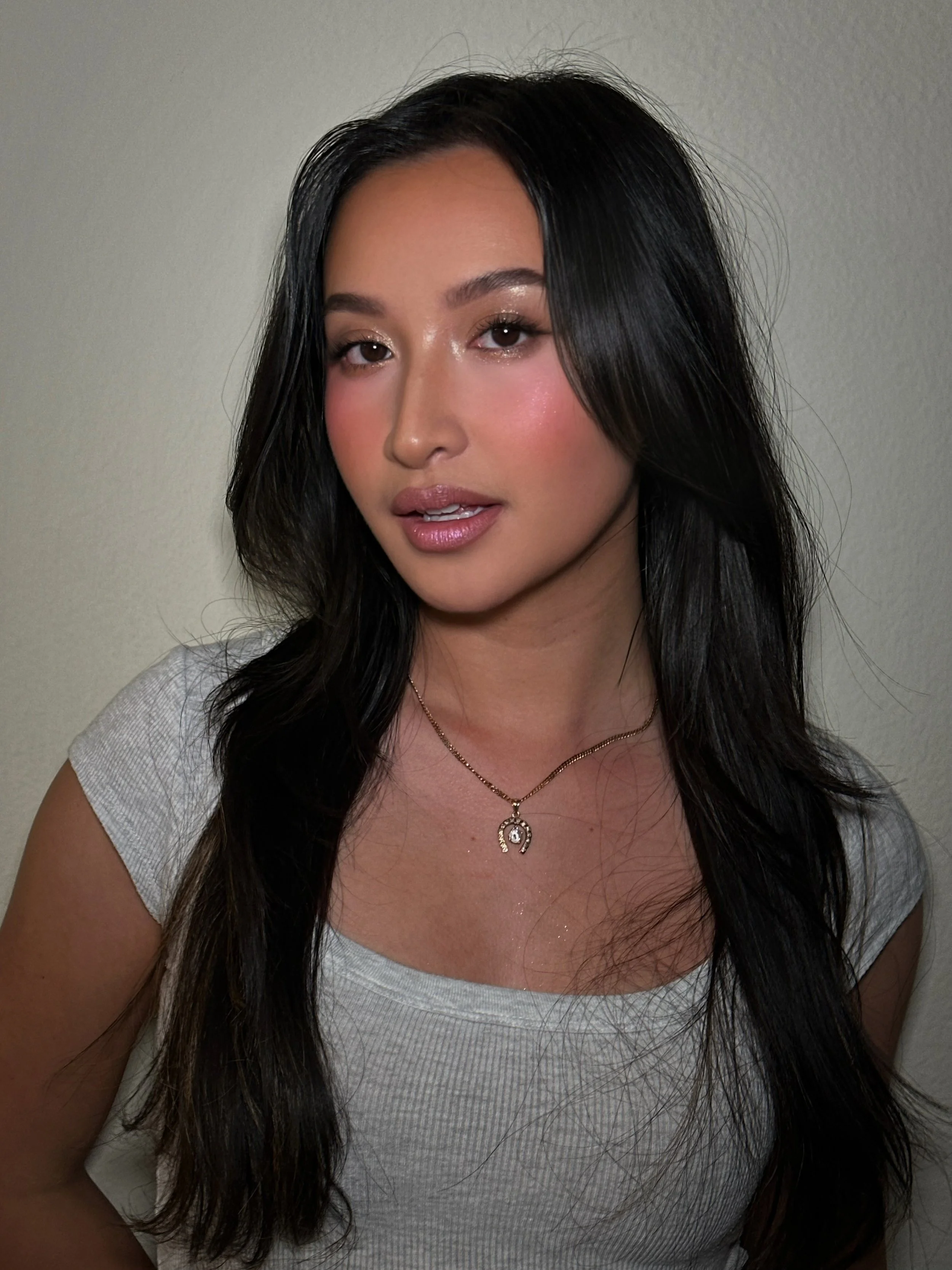 A woman with long black hair, light makeup, and a light gray top, wearing a gold necklace with a pendant, posing against a neutral background.