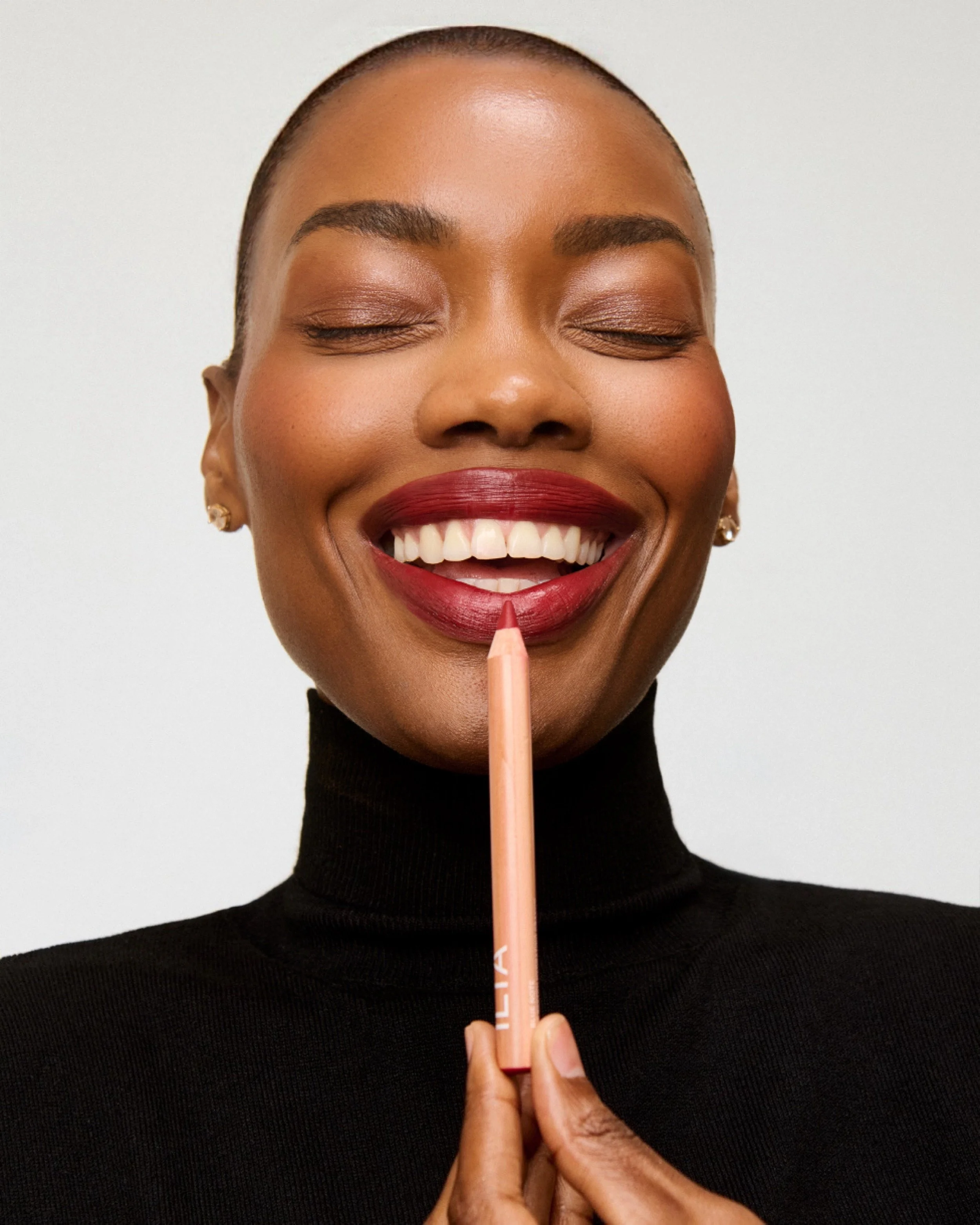 A woman with short hair and dark skin smiling with her eyes closed, holding a lip pencil near her lips.