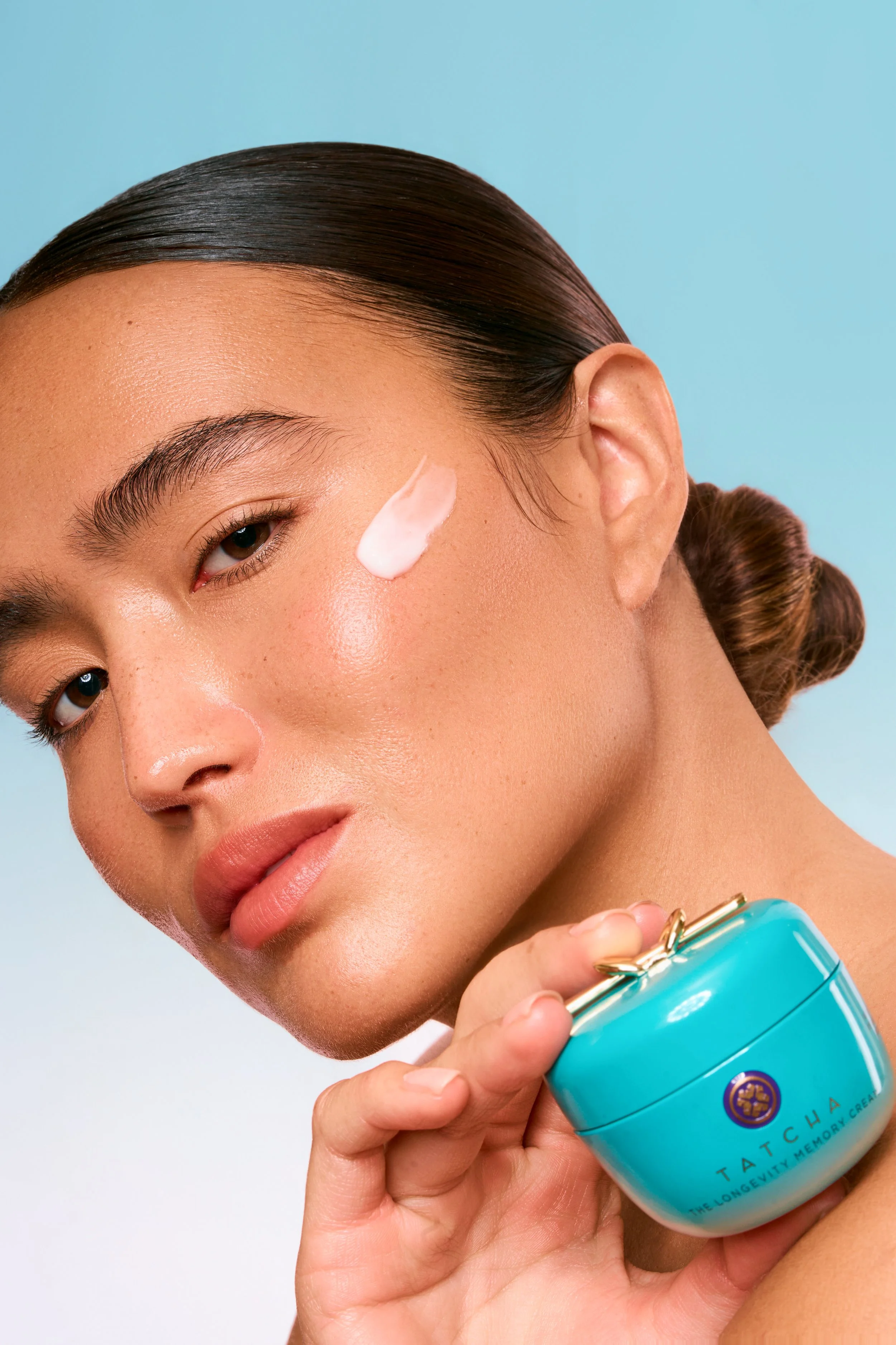 A woman holding a blue jar of Tatcha skincare cream with a small amount of product on her cheek, against a light blue background.