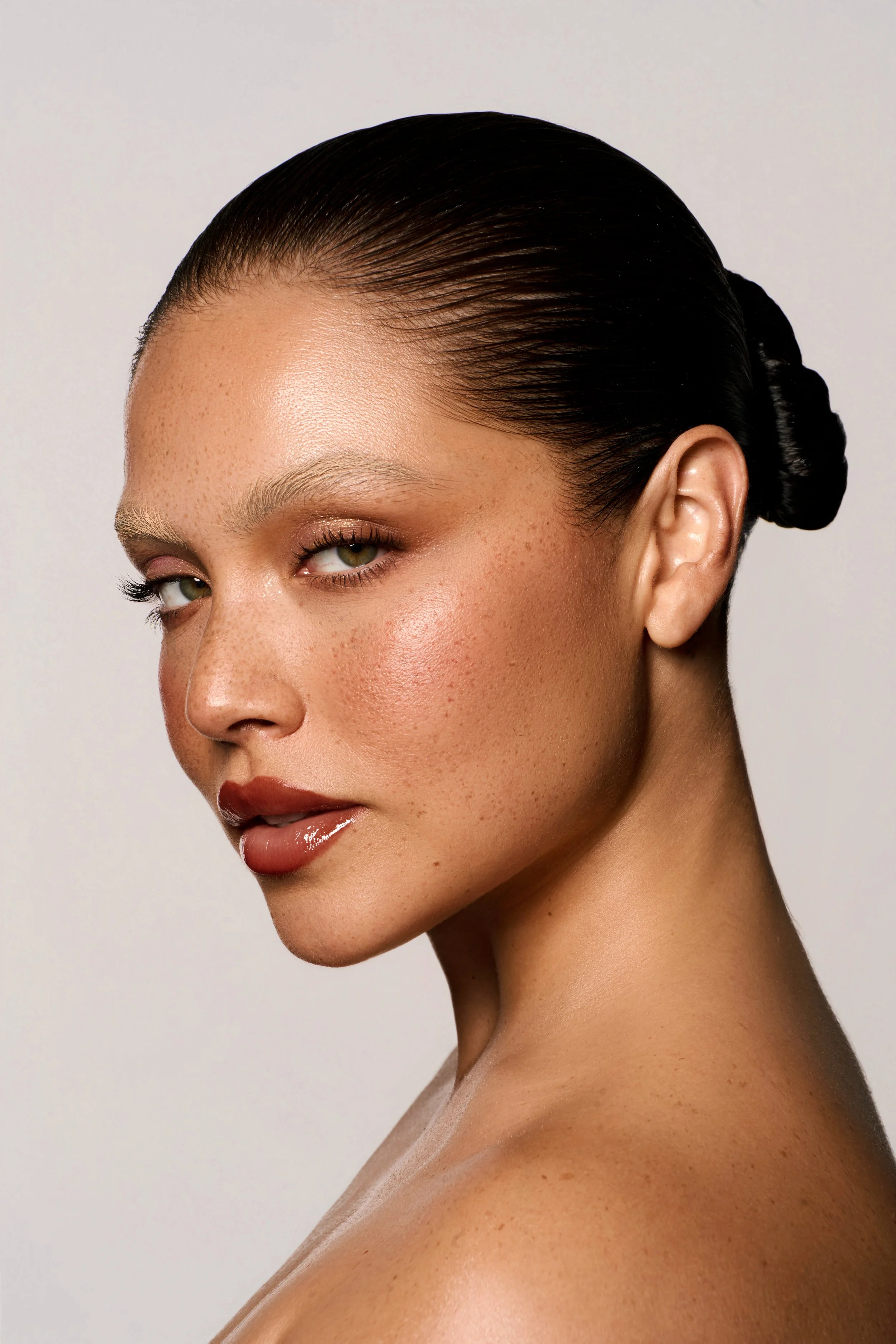 A close-up portrait of a woman with slicked-back dark hair, glowing skin, natural makeup, and wearing nude lipstick, against a neutral background.